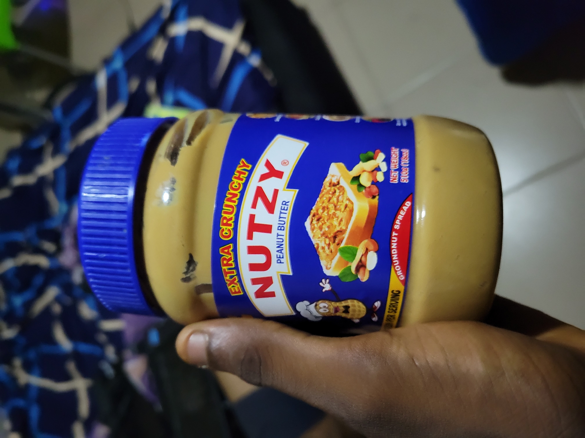 A hand is holding a jar of Nutzy Extra Crunchy Peanut Butter. The jar has a blue lid and a label with a cartoon peanut wearing a chef's hat, a slice of toast with peanut butter, and nuts. The background is a blurry blue and black patterned fabric. The lighting is bright, casting shadows on the jar and hand.