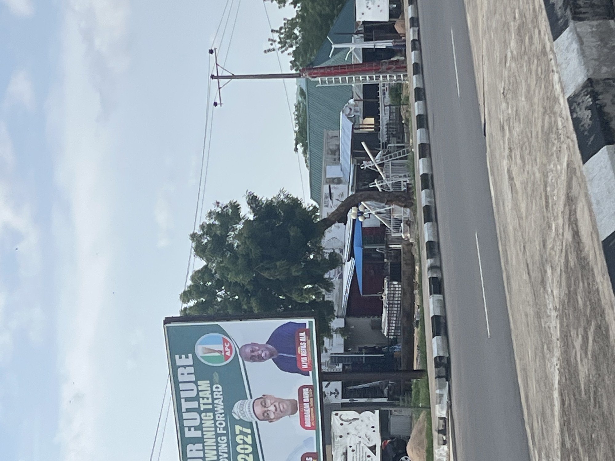 A billboard promoting a political campaign is visible on the left side of the image. The billboard states "OUR FUTURE WINNING TEAM MOVING FORWARD 2027". Two men are pictured on the billboard, with the names "ABUBAKAR BAWA" and "ILIYA KEFAS ALI" below them. The logo for the APC (All Progressives Congress) party is also present. Behind the billboard, a street is lined with buildings and trees. The street has a black and white striped curb. A utility pole with wires is visible on the right side of the image, with a red ladder leaning against it. The sky is overcast. The location is Jalingo, Nigeria.