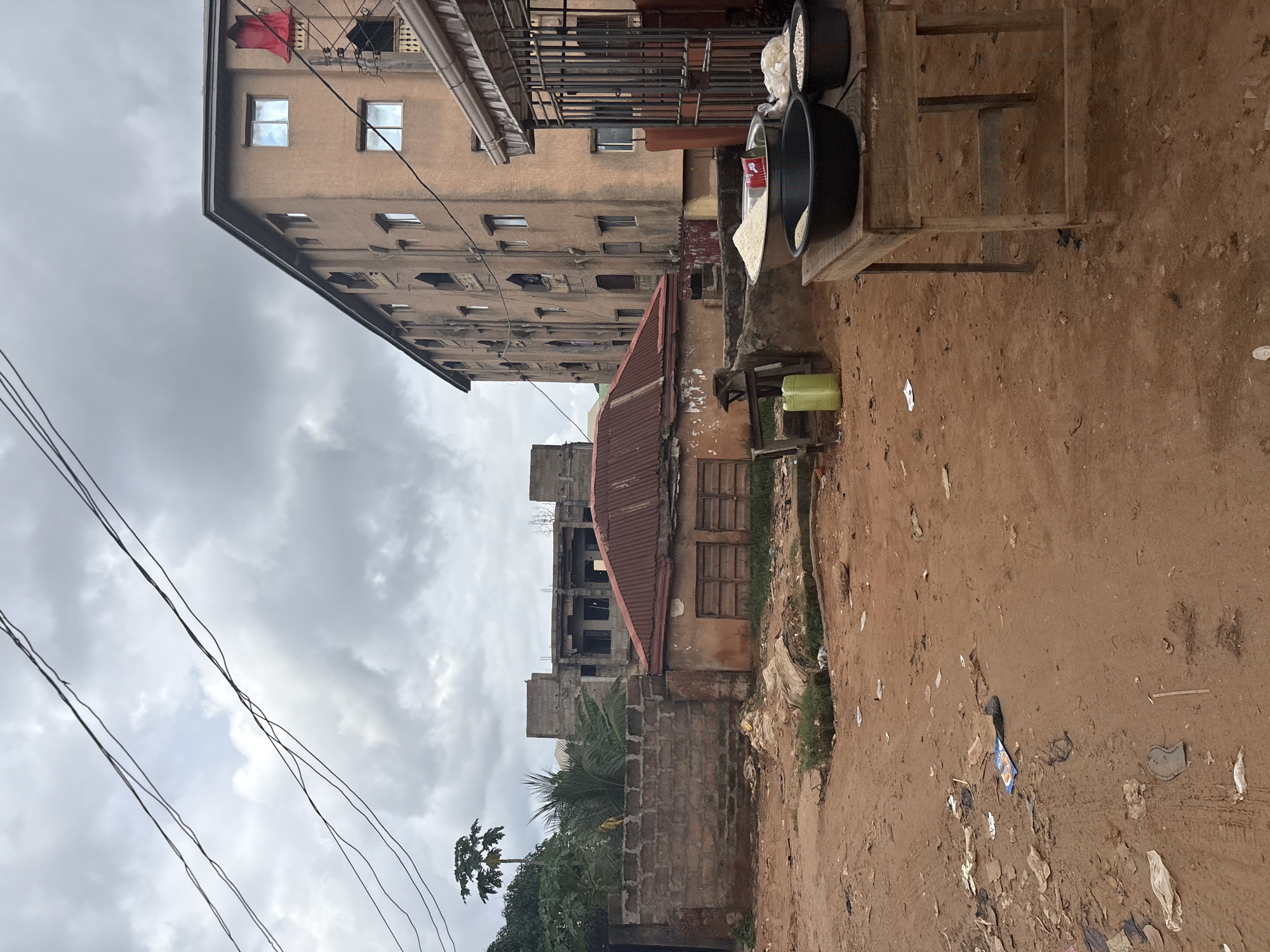 The image depicts an outdoor scene in Nkpor, Nigeria. The dominant subjects are buildings and a dirt road, suggesting an urban or semi-urban environment. A multi-story, light-colored building with many windows dominates the left side of the frame. In front of it, a smaller, older building with a corrugated metal roof is visible. The ground is a reddish-brown dirt road, strewn with litter, including plastic bags and paper scraps.
To the right, a wooden stand holds two large black bowls. One bowl contains a white powdery substance, likely flour or a similar ingredient, while the other contains a lighter-colored granular substance, possibly grains. A red and white tin is also visible on the stand. A green plastic container sits on the ground near the wooden stand.
Further back, behind the smaller building, there are signs of ongoing construction, with exposed concrete structures. Lush green palm fronds and other foliage are visible, indicating a tropical climate. Overhead power lines are strung across the sky, which is overcast and grey, suggesting a cloudy day. The overall impression is one of a lived-in, possibly developing, area with a mix of older and newer structures and the presence of street vending or market activity. There is no discernible text in the image.