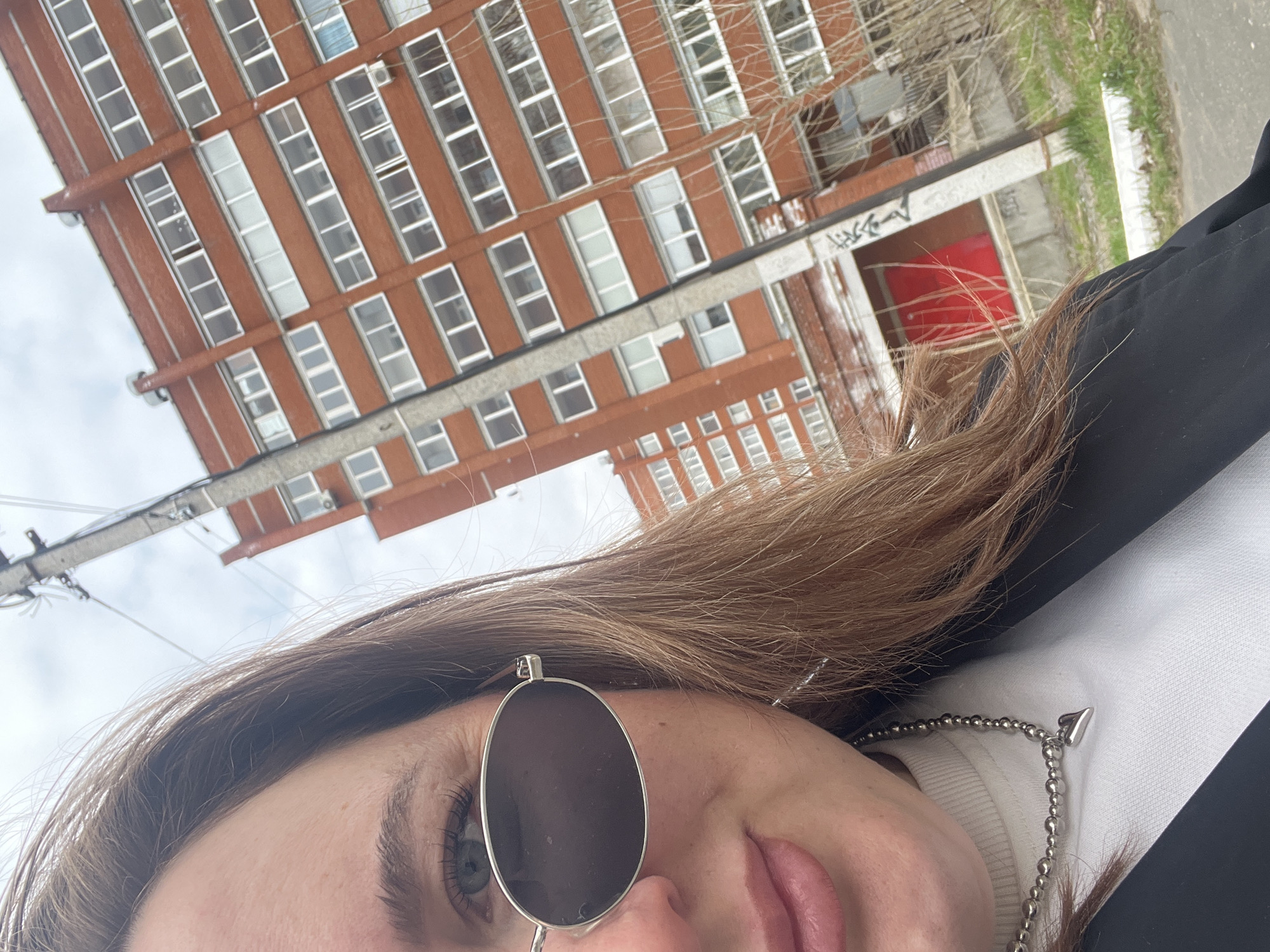 A close-up, slightly tilted selfie photograph shows a woman with light brown hair wearing a round, dark-lensed sunglass over one eye. She has fair skin and is wearing a white turtleneck sweater and a black jacket. A silver ball-chain necklace with an arrow-shaped pendant is visible.
The background features a multi-story brick building with numerous white-framed windows. The building appears to be located in Chaikovsky, Russia. A power line is visible above the building. Another building with a red door and graffiti is partially visible behind the main building. Greenery is visible to the right. The sky is overcast.