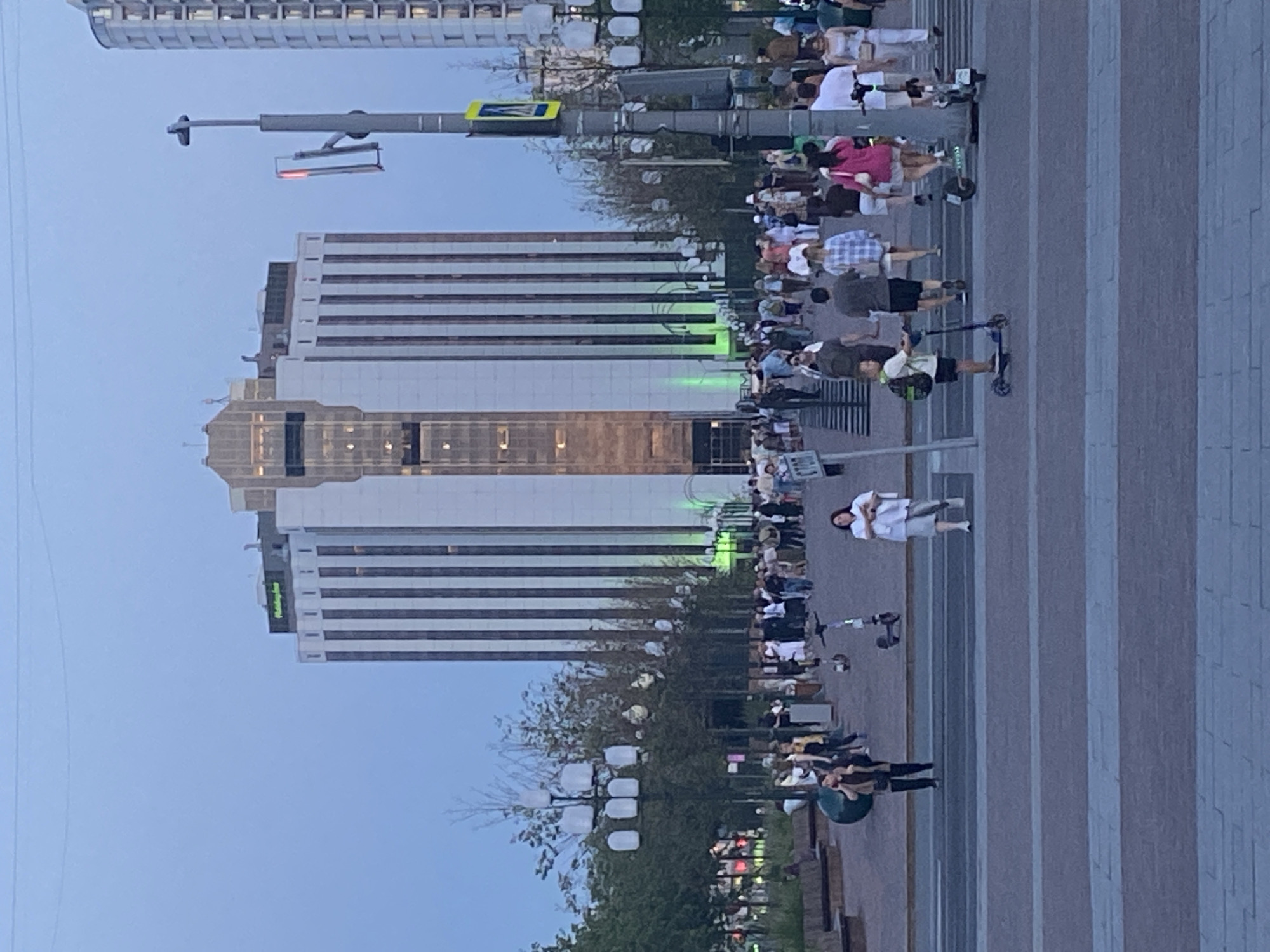 The image shows a wide pedestrianized area in front of a tall, modern building with vertical white and dark elements. The building has a sign at the top that reads "Holiday Inn" in green letters. The sky is overcast and appears to be dusk.
In the foreground, a street with multiple lanes of traffic is visible, with a pedestrian crossing. Several people are crossing the street, and a young boy is riding a scooter. Other people are walking on the sidewalk and in the pedestrian area.
There are streetlights with multiple globes along the sidewalk. Trees and other vegetation are visible behind the people.
The scene appears to be a busy urban area with a mix of pedestrians, vehicles, and modern architecture. The overall atmosphere suggests a public space, possibly a square or plaza, in a city.