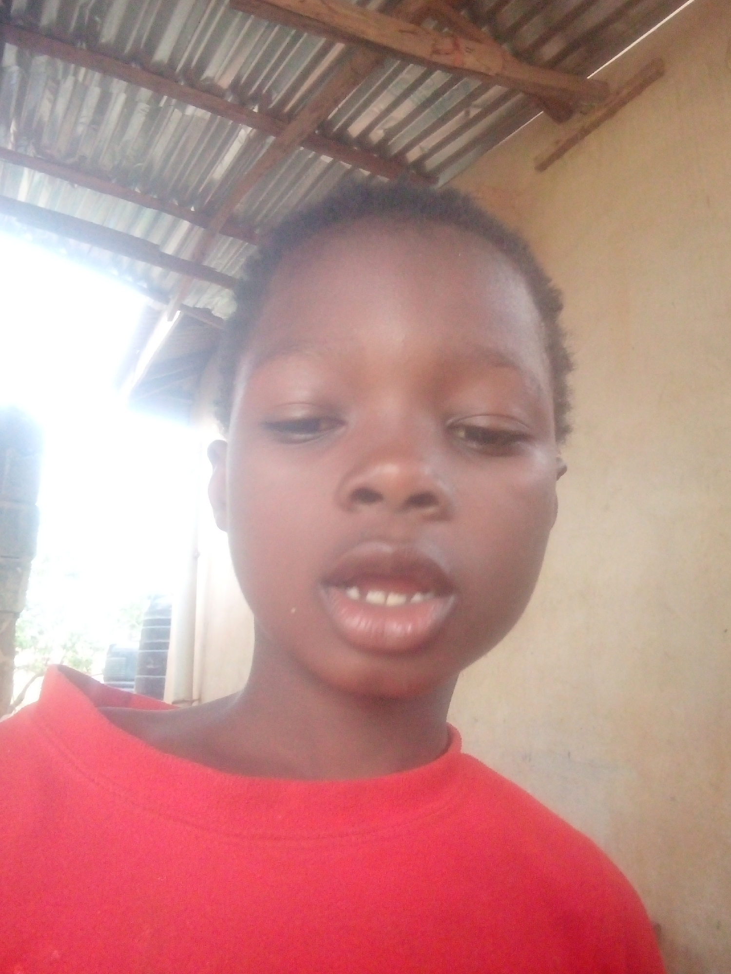 A close-up, eye-level portrait features a young person, likely a child, with dark skin and short dark hair. The individual is positioned centrally in the frame, facing forward with a slightly open mouth, revealing some teeth. Their eyes appear partially closed or are looking downwards. They are wearing a plain red crew-neck shirt.
The lighting is very bright, originating from the left side of the frame, causing significant overexposure and glare on the left side of the subject's face and the background. Overhead, corrugated metal roofing supported by wooden beams is visible, suggesting an outdoor or semi-sheltered setting, possibly a simple structure like a shed or an open-sided building. The bright light on the left also reveals glimpses of what might be foliage or other distant structures. To the right of the subject, the background consists of a plain, light brown wall.
The image captures a moment of the person posing for the photograph. The strong, natural light indicates it is daytime. No specific activities are taking place, and no discernible text is visible.