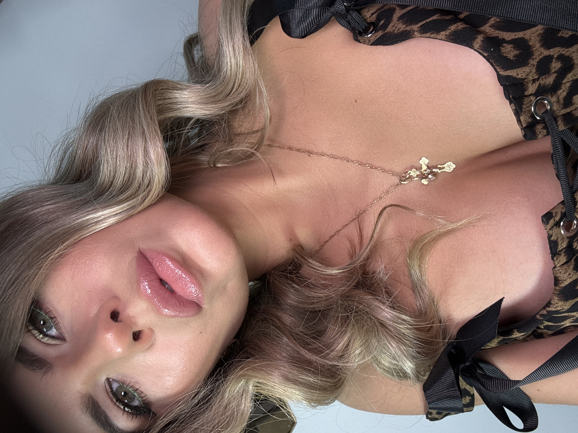 A woman with blonde hair and a leopard print top is positioned in an indoor setting. She is wearing a gold cross necklace and makeup, including eyeliner and lip gloss. There is no information available to determine the location to be in the city of Chaikovsky, Russia.