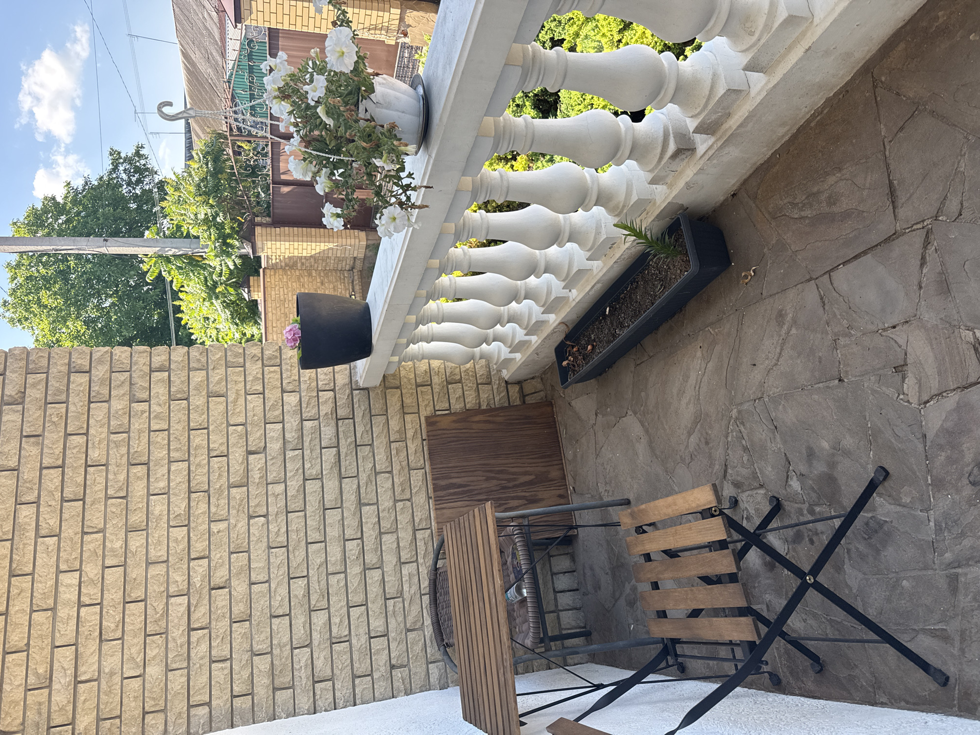 The image depicts an outdoor balcony or patio area in Horbanivka, Ukraine. The setting is a sunlit day, as indicated by the bright sky and strong shadows. The balcony is furnished with a round wooden table and two folding chairs, suggesting a place for outdoor dining or relaxation.
The balcony features a white decorative balustrade with ornate spindles. White petunias are visible in a white pot hanging from the railing, adding a touch of color and life to the scene. Next to the balustrade, a black planter box contains a small green plant. The floor of the balcony is paved with large, irregularly shaped stone tiles.
To the left of the balcony, a light-colored brick wall rises, forming part of a building. Green trees and foliage are visible beyond the balcony railing, suggesting a residential or garden area. Overhead, power lines and a cloudy blue sky are present, with a hook or bracket attached to a pole.
The overall impression is one of a peaceful, outdoor space ready for use, perhaps enjoyed on a warm day in Ukraine. No people are visible in the image, and there is no visible text.