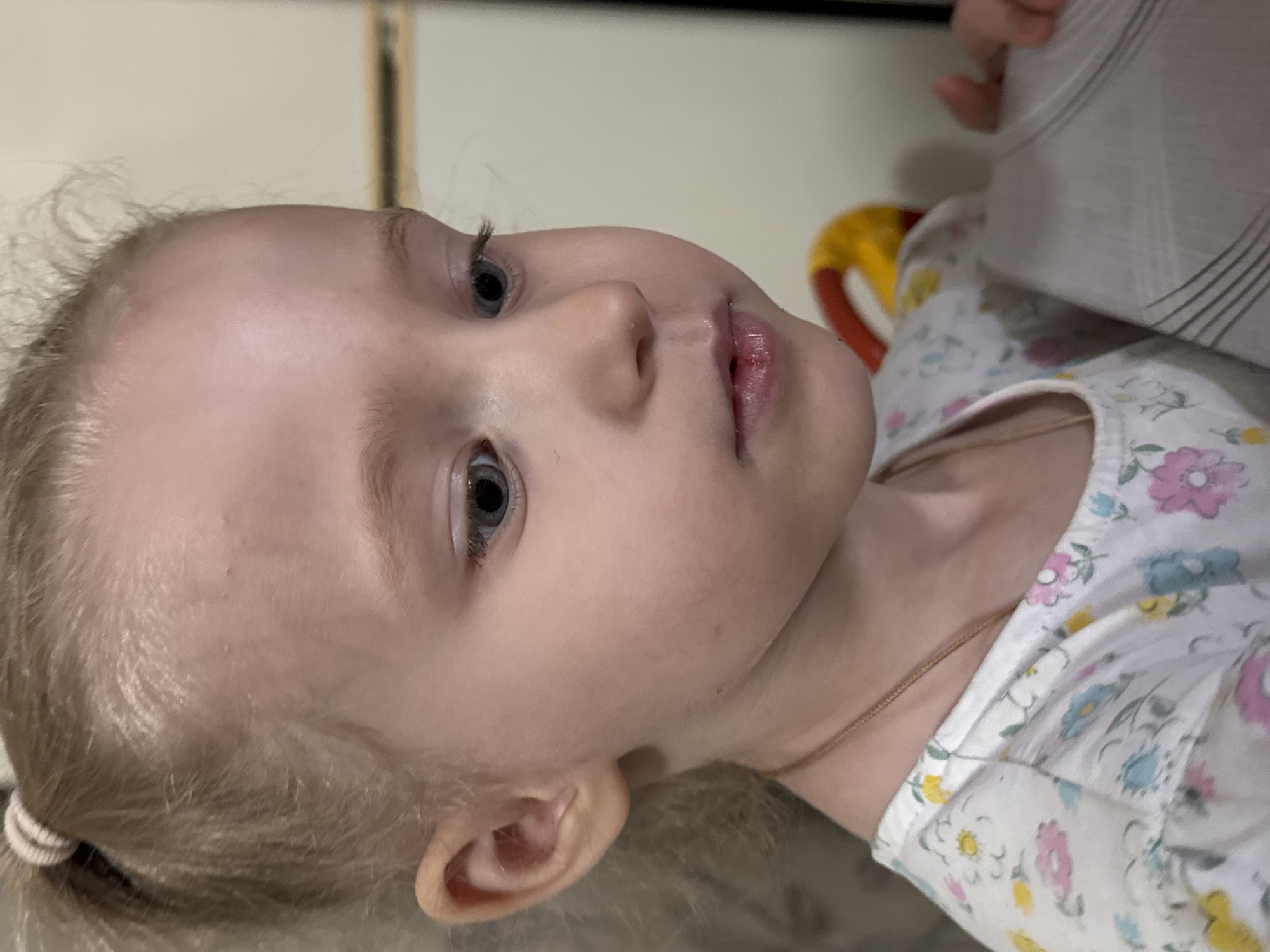 A close-up, slightly rotated image shows a young child with fair skin and light blonde hair pulled back into a small ponytail with a pale rubber band. The child has gray-blue eyes and is looking upward. The child is wearing a light-colored floral print shirt with pink, blue, and yellow flowers. A thin gold chain is visible around their neck. Behind the child are indistinct shapes and colors, including a partial view of a yellow object and a striped, light-colored fabric or surface. The environment appears to be indoors. There is no indication of a location within Nizhnyaya Gar', Russia from the image.