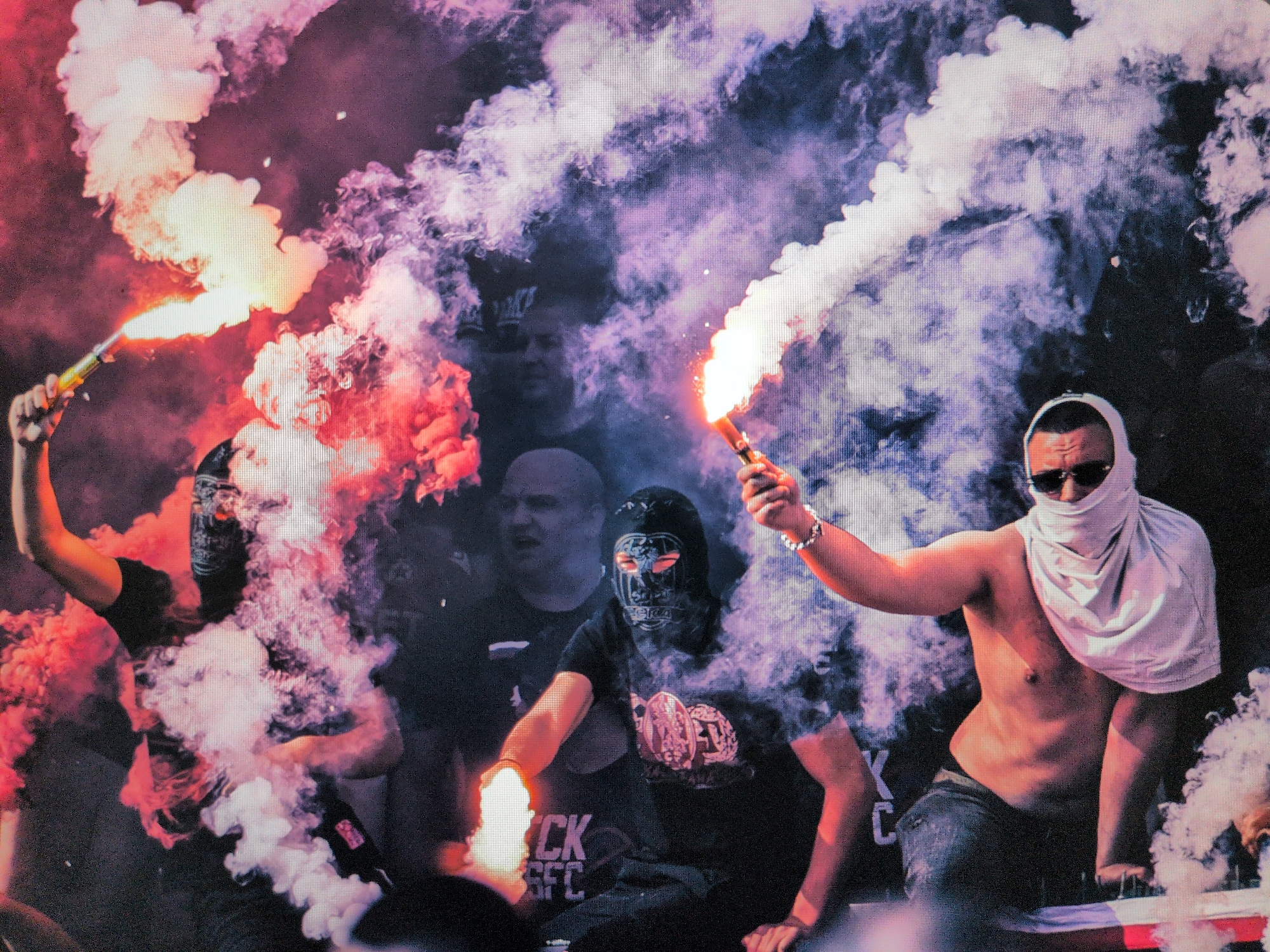A dynamic, low-light image captures a scene of fervent activity, likely during an evening or nighttime event, characterized by dense, colorful smoke and the illumination of flares. In the foreground, three individuals are prominently featured amidst clouds of red, white, and purple smoke.
To the left, a person wearing a black mask or balaclava and a dark long-sleeved shirt holds an ignited red flare, its bright flame and thick smoke rising upwards. In the center, another individual in a black t-shirt and a similar black mask kneels or sits, holding a red flare that emits a fiery glow and dense smoke. Partial text "CK SFC" is visible on their shirt, along with a circular emblem design. To the right, a shirtless person in jeans, with a white cloth wrapped around their head and face, creating a makeshift mask, holds a white or orange flare. They wear sunglasses and a silver bracelet on their wrist.
The flares provide the primary light source, casting dramatic shadows and highlighting the billowing smoke, which dominates the mid-ground and background. Behind the main subjects, several other indistinct figures are partially visible through the smoke, suggesting a crowd or group gathering. The dark, obscured background implies an undefined setting, possibly a stadium, street, or event venue. The atmosphere conveyed is intense, passionate, and perhaps rebellious or celebratory, common in fan culture, protests, or public demonstrations.