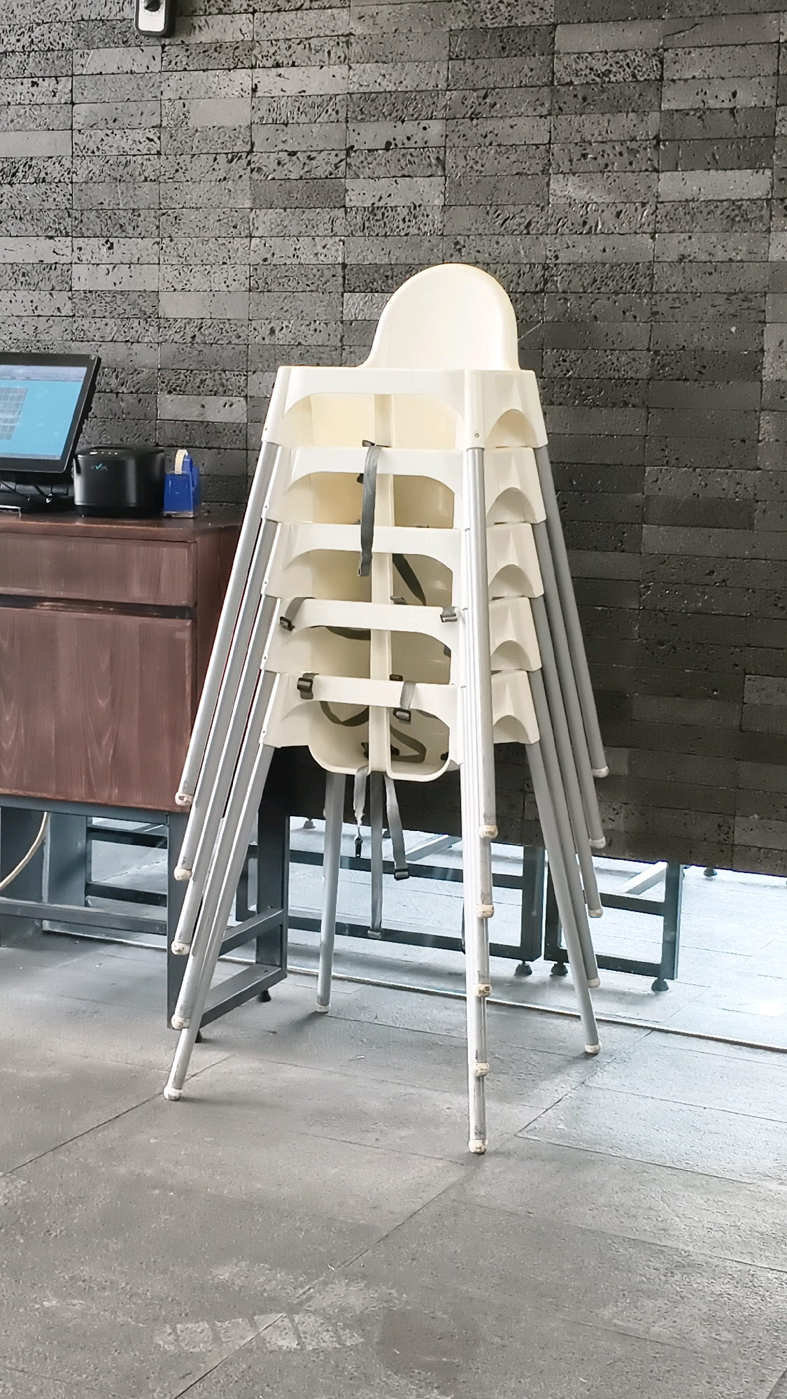 A stack of white plastic high chairs with gray metal legs is centrally positioned in the foreground of the image. The high chairs are neatly nested, with visible straps on each one. To the left of the high chairs, a dark brown wooden cabinet with a metal frame is visible. On top of the cabinet, a black electronic device and a blue tape dispenser can be seen. A computer monitor displaying a blue screen is also on the cabinet, slightly angled. The background features a textured gray brick wall. The floor in the foreground is made of gray concrete tiles, with some lighter patches indicating wear or debris. The environment appears to be indoors, possibly a restaurant or a public space in Bekasi, Indonesia, given the context provided. There are no people visible in the image.