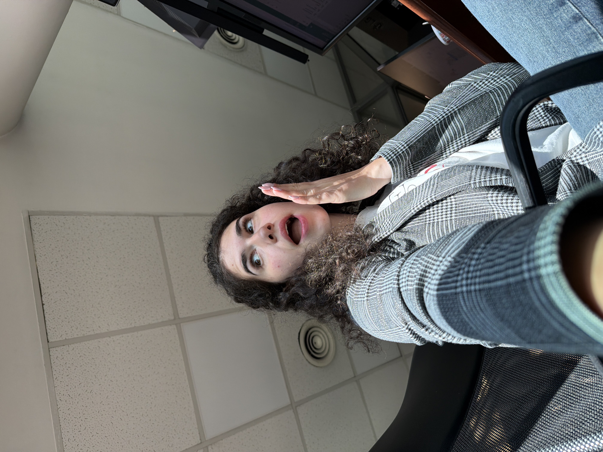 In Sofia, Bulgaria, a young woman with curly dark hair sits in an office chair, looking upwards and to the right with a surprised expression. Her mouth is open, and her right hand is raised to her cheek, palm facing outward. She is wearing a grey plaid blazer over a white t-shirt and blue jeans. The background shows a suspended tile ceiling with a circular air vent and a glimpse of a computer monitor on the right. The lighting suggests it is daytime, with sunlight casting shadows.