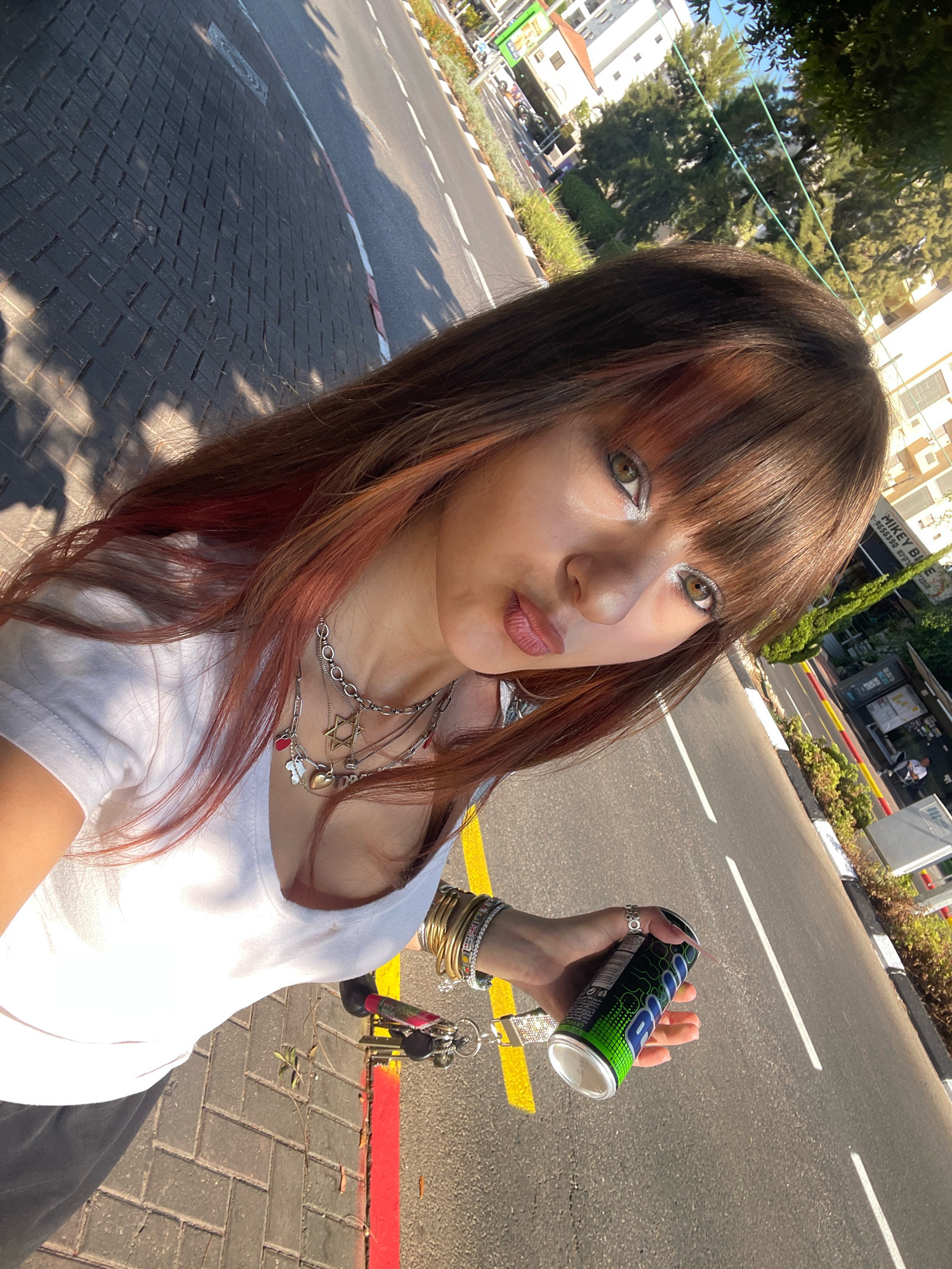A young woman with long, brown hair with reddish highlights stands on a paved sidewalk next to a street in Raanana, Israel. She is wearing a white t-shirt and several necklaces. In her right hand, she holds a green and black can of an energy drink. The background shows a street with cars, buildings, and trees. The sun is casting shadows on the sidewalk.