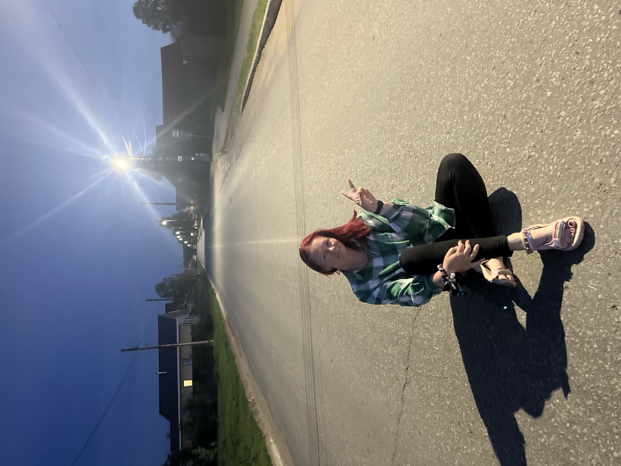 A woman sits cross-legged on an asphalt road in what appears to be the city of Chaikovsky, Russia. She wears black leggings, a green and white plaid shirt, and sandals. She is gesturing with her left hand. The photo appears to be taken at dusk or dawn, with a street light visible in the distance. Buildings and vegetation line the sides of the road.