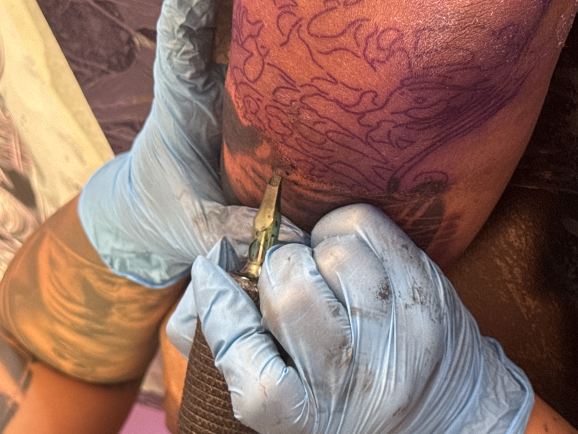 The image depicts a close-up view of a tattoo being applied to a person's leg. The tattoo artist, wearing blue nitrile gloves, is holding a tattoo machine and working on a design marked with purple stencil ink on the skin. The skin appears to be of darker complexion, and the background is out of focus, showing a reddish-brown textured surface and some fabric. The lighting suggests an indoor setting, possibly a tattoo studio. The scene is focused on the action of tattooing, with no discernible location in Vigo, Spain.