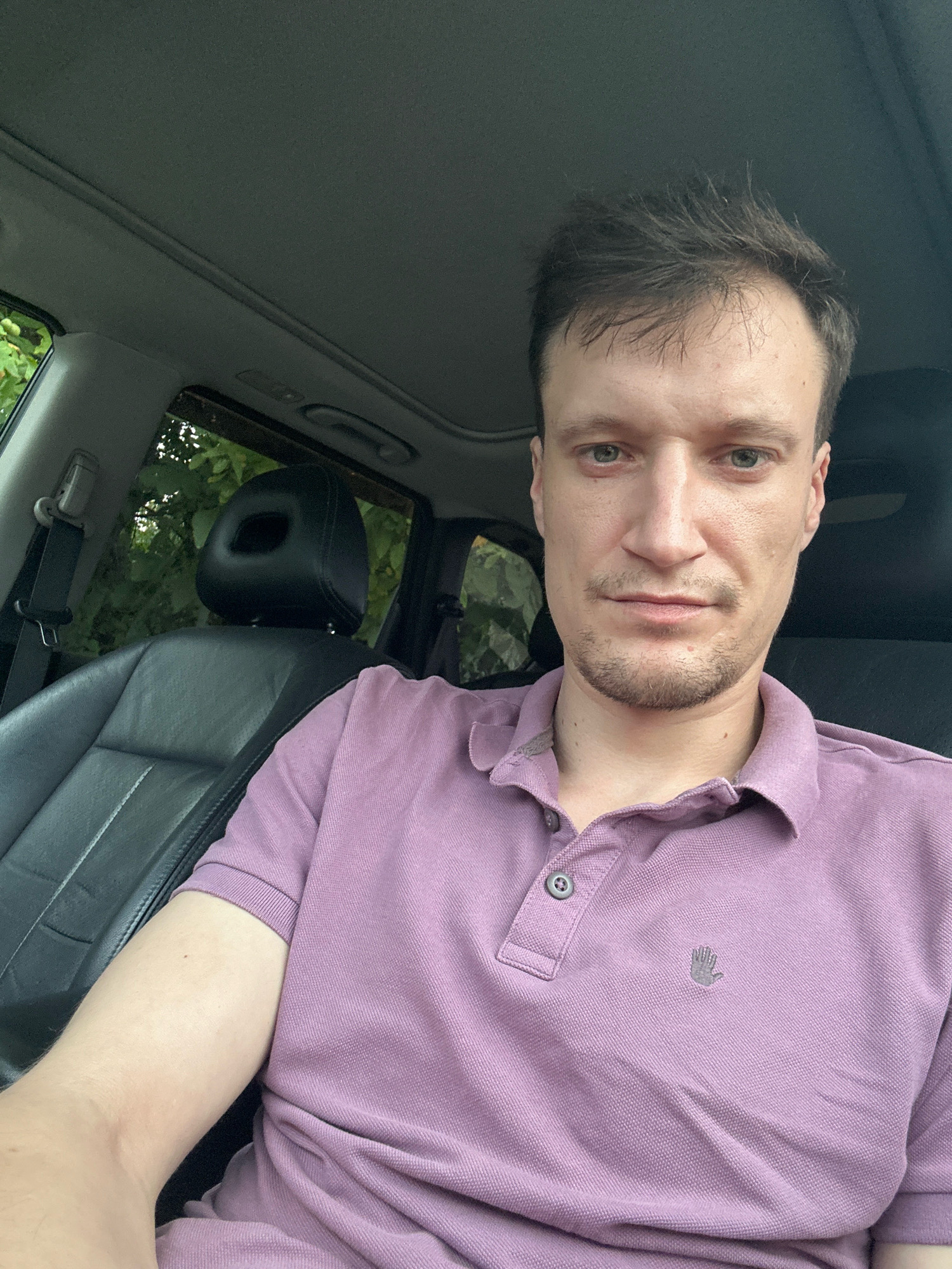 This is a close-up, selfie-style photograph taken inside a car. The subject is a fair-skinned man with short, dark brown hair and a light stubble on his chin. He is wearing a light purple polo shirt with a small, grey embroidered hand logo on the left chest. The interior of the car is visible, including black leather seats, a seatbelt, and a sunroof. Through the car window, lush green foliage can be seen, suggesting an outdoor setting. The man is looking directly at the camera with a neutral expression. The image is well-lit, possibly from natural daylight. The location of Shatava, Ukraine is not discernible from the visual elements in the photograph.