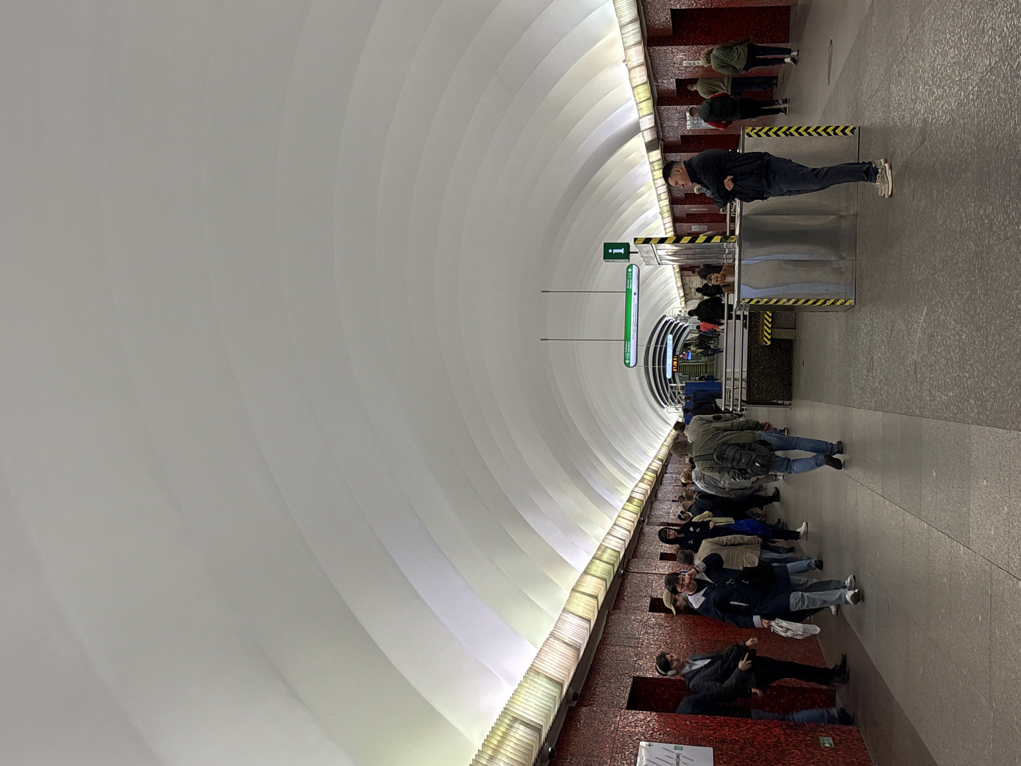The photograph depicts a scene within the Avtovo metro station in Saint Petersburg, Russia. This station is known for its elaborate architectural design, particularly the high, arched ceiling constructed with ribbed, white panels.
Several individuals are visible in the image. On the right side, near a set of turnstiles with yellow and black hazard stripes, a person in a dark jacket and jeans leans against the turnstile. Other individuals pass through the turnstiles. Along the right edge of the image, people are standing in line, some facing the camera, while others are oriented away from it. The station walls are decorated with red, textured material. A green directional sign with an 'i' is suspended from the ceiling.
The perspective is taken from a low angle, emphasizing the station's high, curved ceiling. The floor is covered with light-colored tiles. The overall impression is a busy, functional space with a strong architectural character.