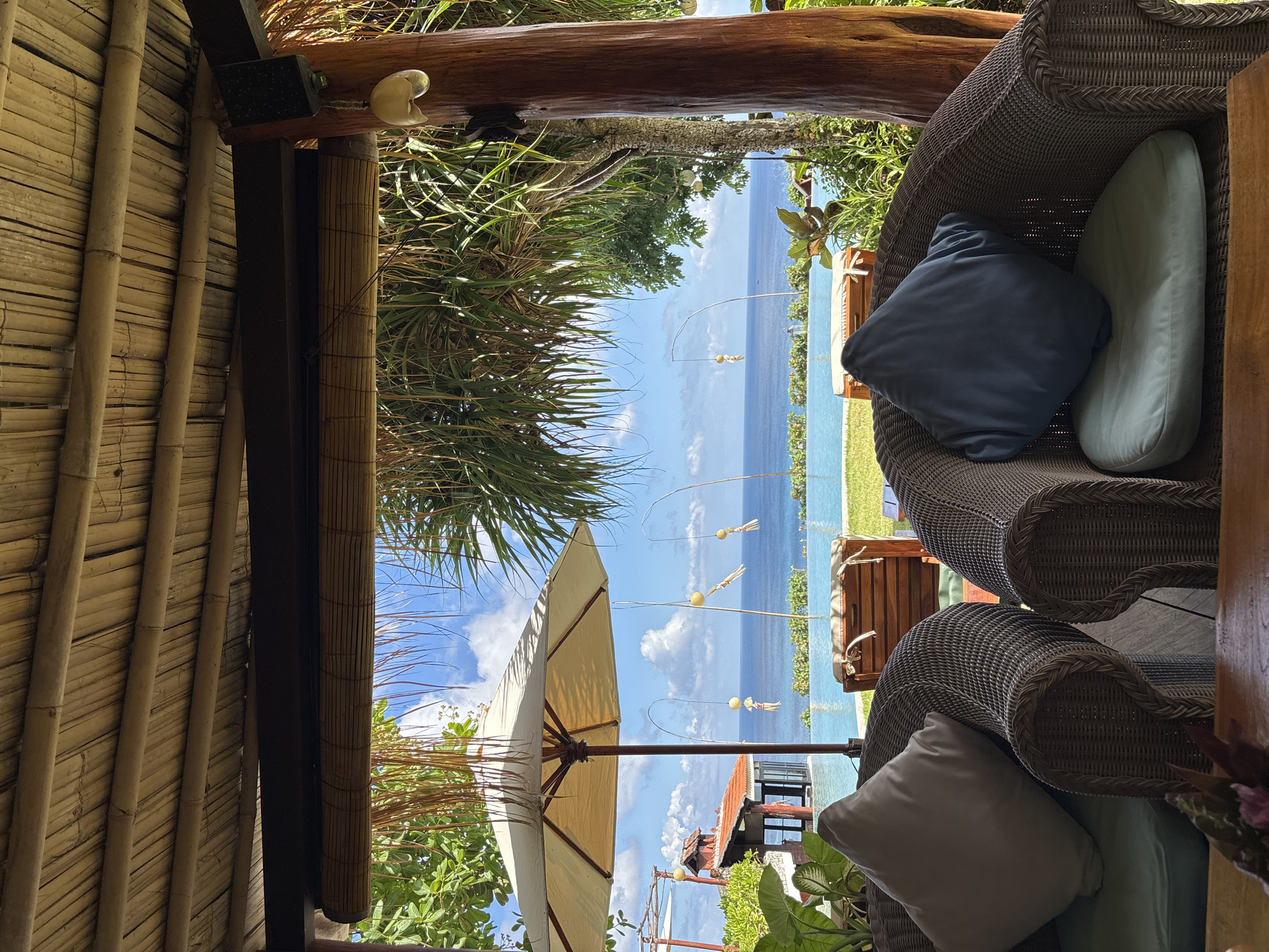 View from a covered patio in Kuta Selatan, Indonesia, with woven bamboo walls and ceiling. Two wicker chairs with cushions are visible. The patio offers a view of the ocean, vegetation, an umbrella, and a building with a red roof.