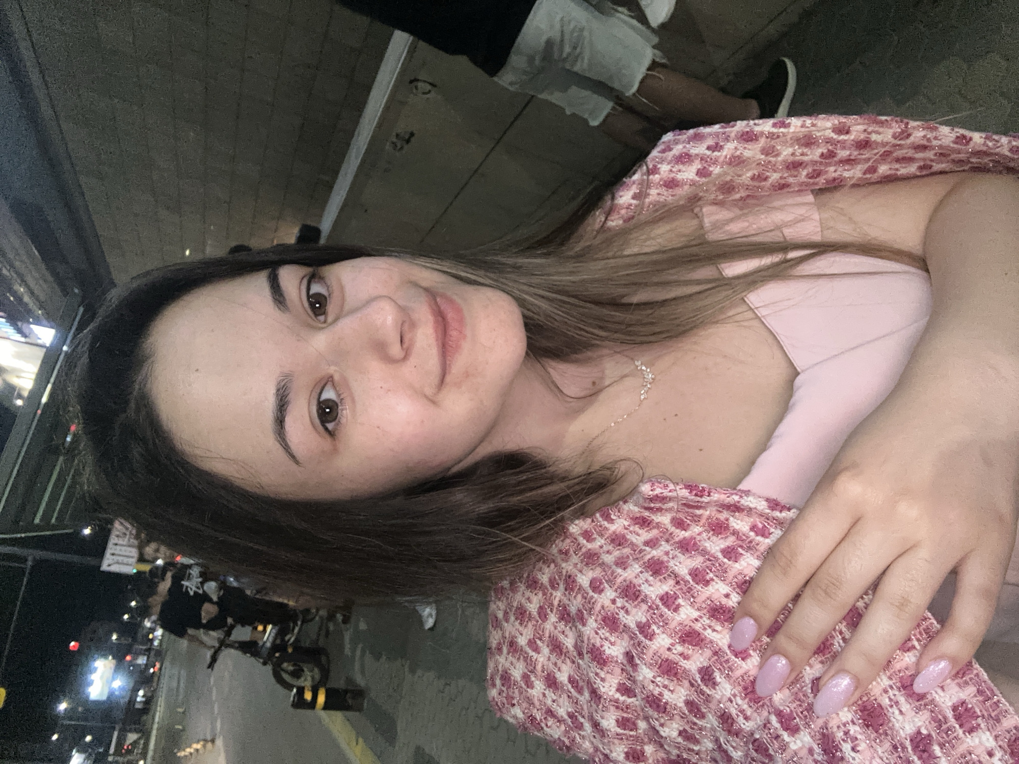 A young woman, approximately in her twenties, is centered in a selfie taken outdoors at night on a city street in Sofia, Bulgaria. She has long, dark brown hair, light brown eyes, and a slight smile while looking directly at the camera. Visible freckles dot her skin. She wears a light pink, possibly strapless or off-the-shoulder top, under a pink and white textured, tweed-style jacket draped over her shoulders. A delicate silver-toned necklace with small clear stones adorns her neck. Her left hand is raised to her chest area, revealing oval-shaped nails painted with a shimmering light pink or lavender polish.
To the left of the subject, a paved sidewalk with black and yellow bollards leads to a street illuminated by multiple distant streetlights, causing lens flares. A person in a black t-shirt with a white graphic (possibly a "Stussy" logo) and dark pants stands next to a small electric scooter or bicycle. Another individual is partially visible behind them. Further down the street, blurred city lights and a red traffic signal are observable.
The background on the right consists of a dark grey, horizontally textured concrete wall, likely part of an urban structure or underpass. In the lower right corner, the legs of another person are visible, wearing light-colored denim shorts with frayed hems and dark athletic shoes with white soles. The overall scene depicts an evening urban setting.