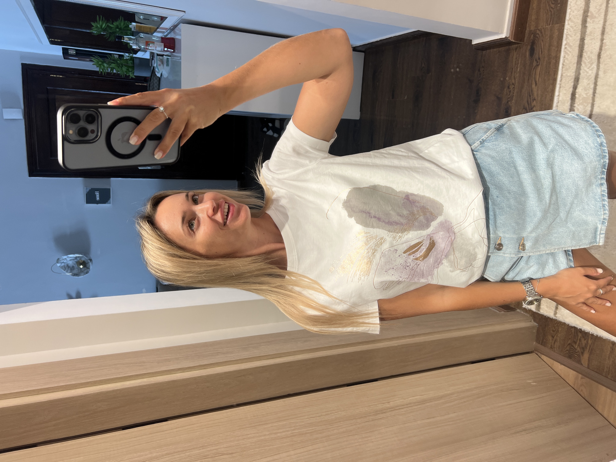A blonde woman takes a selfie in a mirror, her phone held in her right hand. She is wearing a white t-shirt with a watercolor-style print and a light blue denim skirt. The background appears to be a hallway or dressing area, with a wooden door on the left and a dresser or cabinet on the right. There is a framed picture on the wall and some bottles on the dresser. The flooring is dark wood with a rug visible. The overall impression is a casual indoor setting, possibly in an apartment or hotel room in Sharm El Sheikh, Egypt.