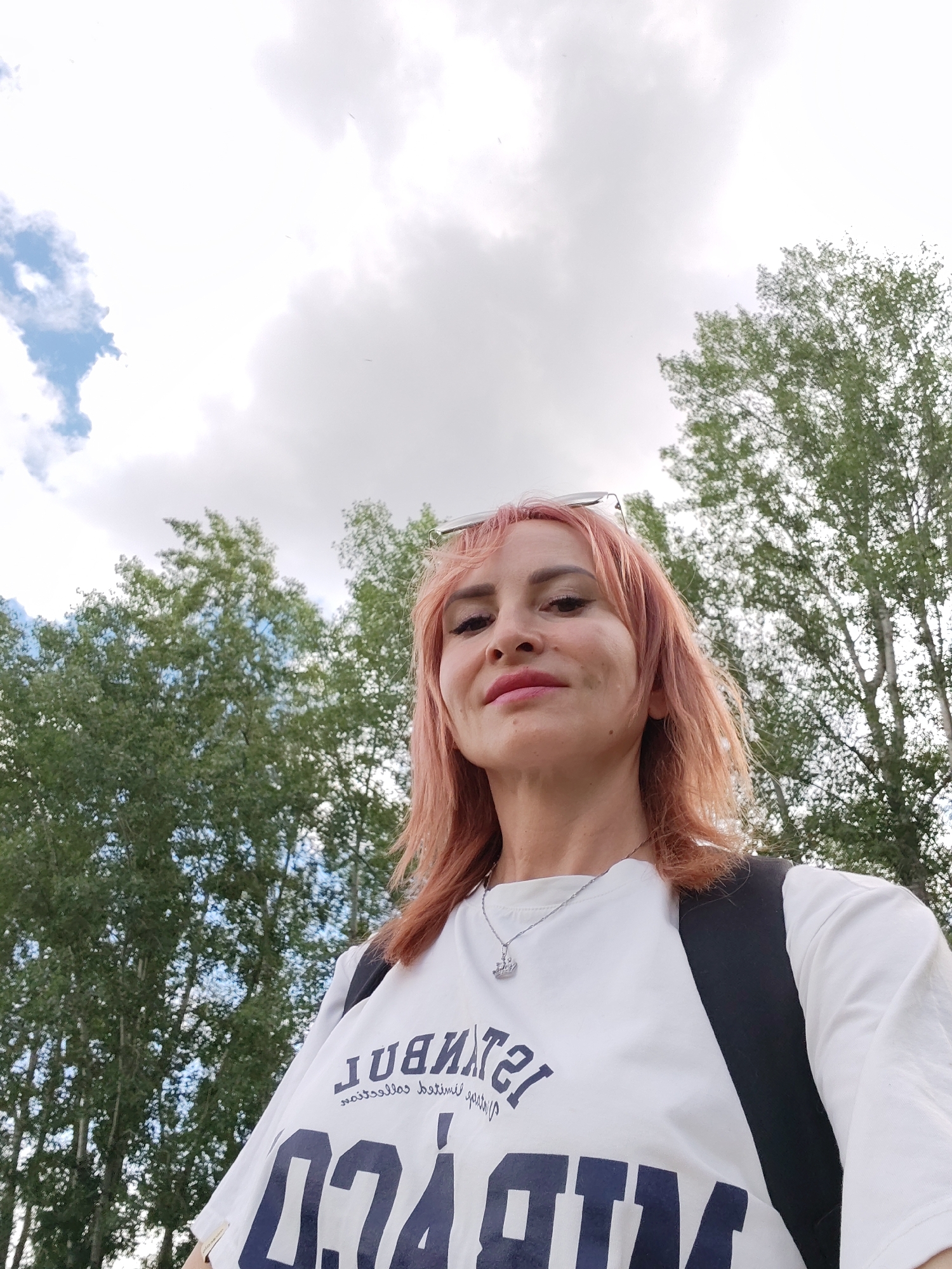 This is a medium shot, selfie-style photo of a woman with light pink hair, looking towards the camera with a slight smile. She is outdoors, with a backdrop of trees and a cloudy sky. She is wearing a white t-shirt with dark blue text and graphics. A silver necklace with a small pendant is visible around her neck. She also appears to be wearing sunglasses pushed up onto her head, and has a black backpack on. The lighting suggests it is daytime, and the sky is overcast with patches of blue sky visible. The presence of deciduous trees indicates it is likely spring or summer. The text on her shirt includes what appears to be "ISTAMBUL" and some other graphics. The location context provided is Chaikovsky, Russia.