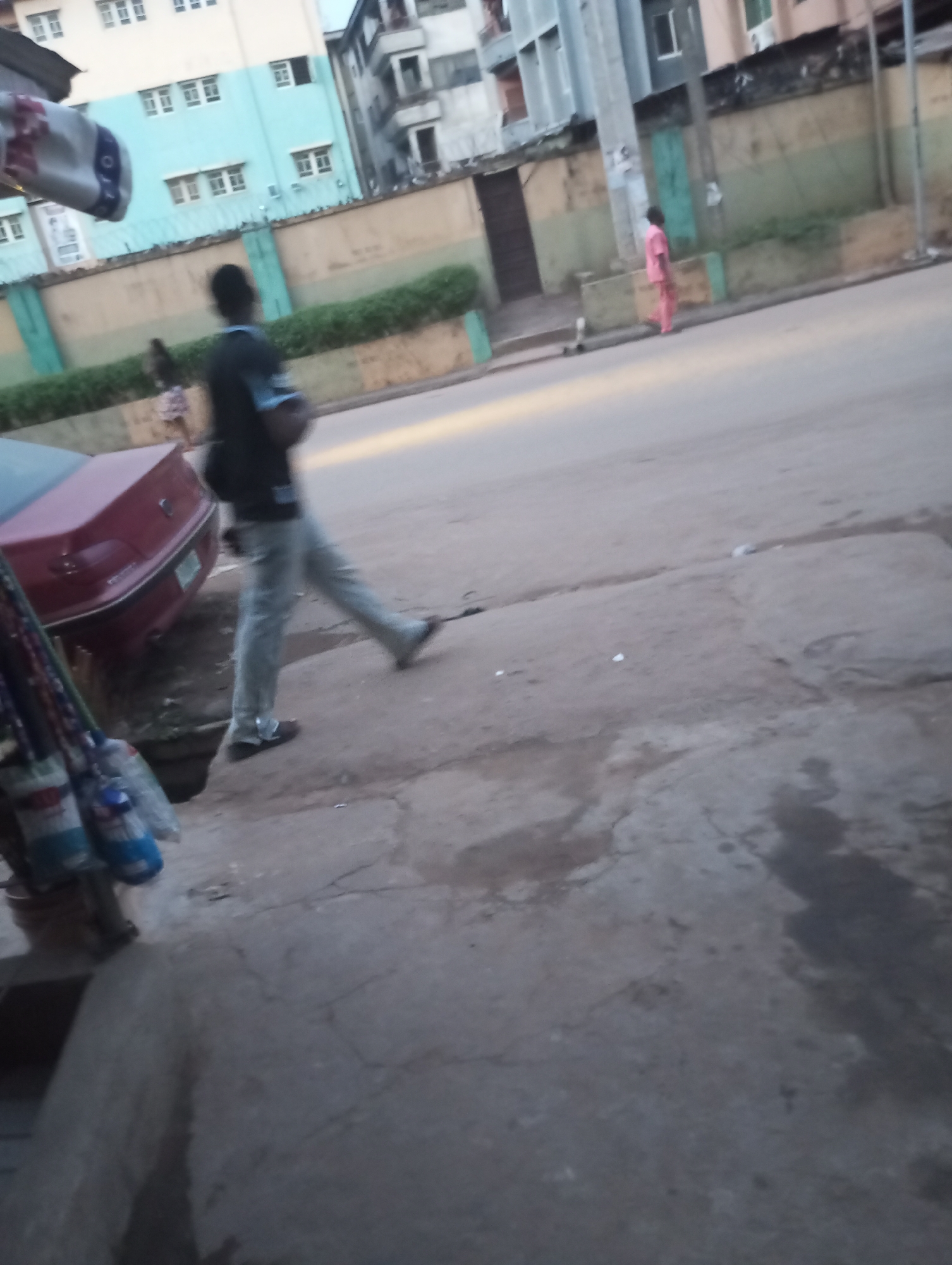 The image presents a slightly blurred, eye-level view of an urban street scene, likely captured in Onitsha, Nigeria, during late afternoon or early evening due to the soft, natural lighting.
In the foreground on the left, various items, including bags and bottles, hang from a display, mostly out of frame. One white bag has an indistinct blue pattern and illegible text. Adjacent to this, the rear of a red sedan is parked. A concrete sidewalk extends from the foreground on the right, meeting a paved road that stretches into the distance.
Several people are visible in motion, contributing to the overall blurriness of the scene. In the mid-ground, a person wearing a dark shirt and light-colored trousers is captured mid-stride, walking from right to left. Further down the street on the right, another individual wearing a light, possibly pink, full-body outfit walks away from the camera. A third, faint figure can be seen in the distance on the left, near a building.
The background features a mix of buildings and street infrastructure. On the left, a multi-story building with light blue and white walls and numerous windows stands behind a low wall, painted in alternating tan and green sections, topped with fencing and barbed wire. Some green foliage is also present above this wall. On the right side of the background, an older, taller building with visible balconies and a muted greyish facade is visible. The scene depicts a typical moment of activity on an urban street. No legible text is visible in the image.