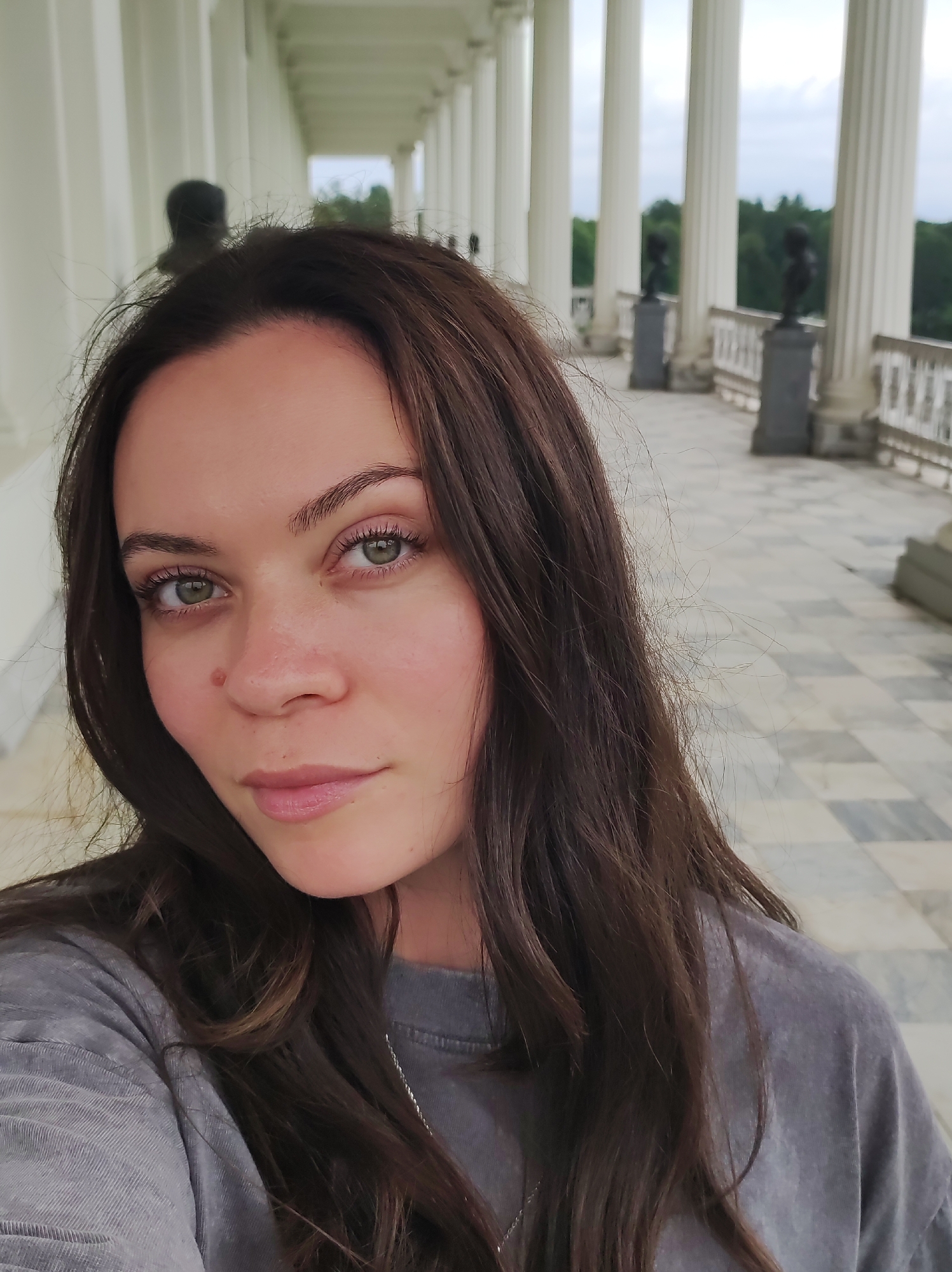 A close-up selfie shows a young woman with long, dark brown hair framing her face. She has green eyes, fair skin with a subtle blush, and a small mole on her right cheek. She is wearing a gray t-shirt and a thin silver necklace. The background is a wide, covered walkway with a colonnade of white pillars. A black railing runs along the edge of the walkway, and beyond it, a checkered tiled floor is visible. In the distance, there are busts on pedestals and lush green trees. The sky is overcast.