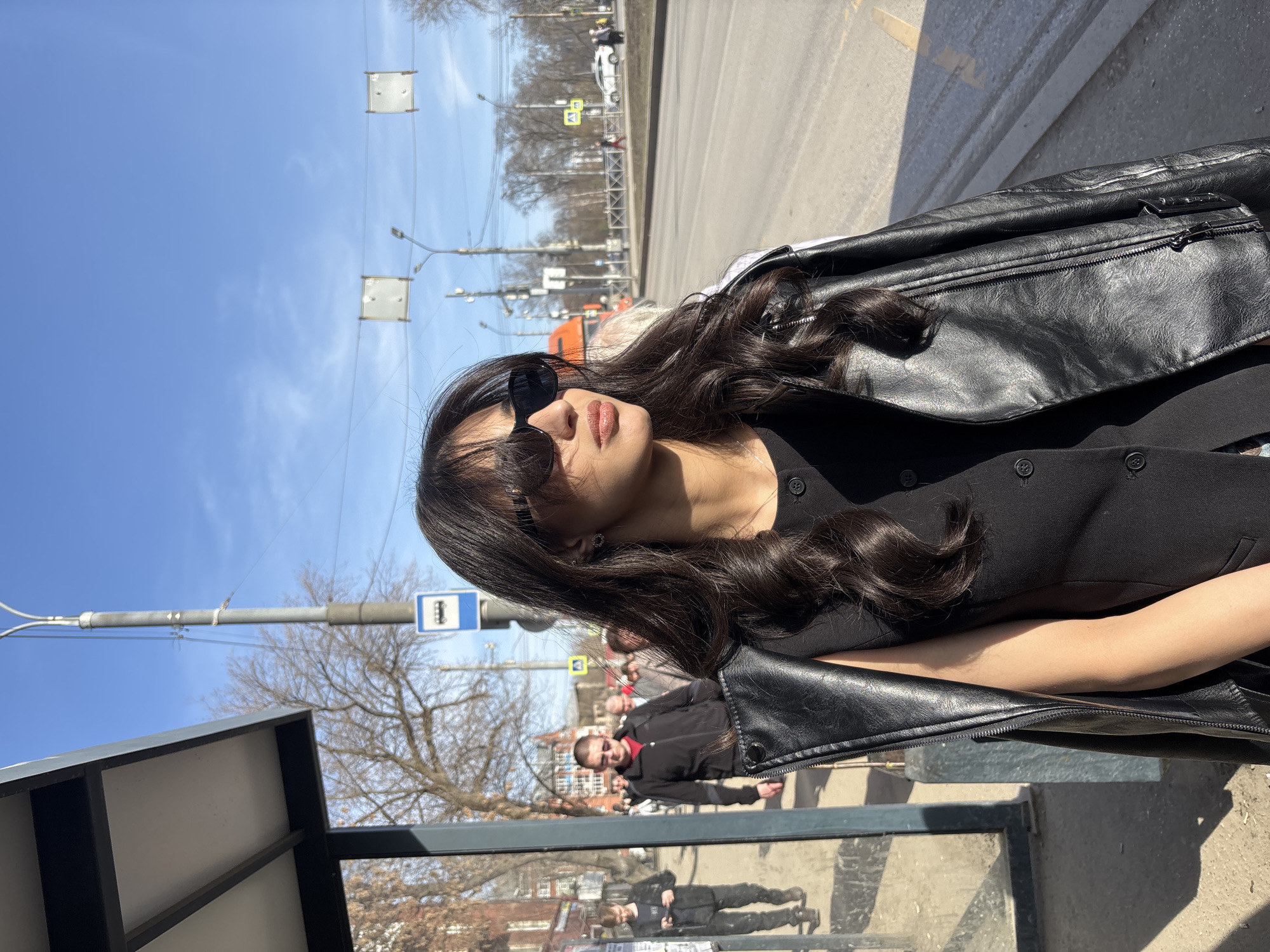 The image depicts a woman with long, dark hair and sunglasses standing at what appears to be a bus stop. She is wearing a black outfit and a black leather jacket. In the background, there is a street with a bus stop sign. People can be seen walking on the sidewalk, and there are buildings visible in the distance. The city is likely Perm, Russia. The sky is blue. The woman is looking away from the camera.