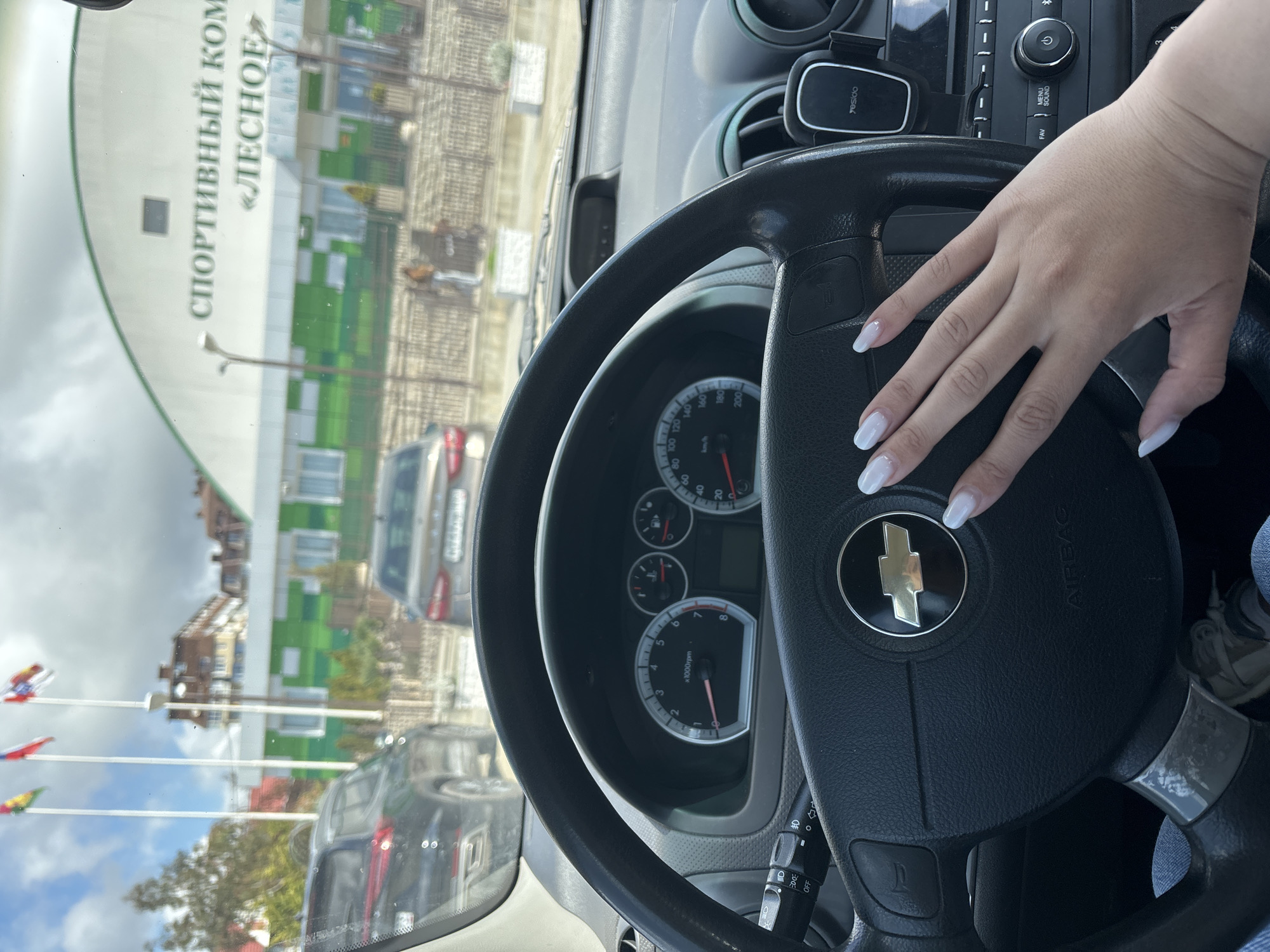 The image, taken from the driver's seat of a car, shows a woman's hand with white nails resting on the steering wheel, which features a Chevrolet logo. The dashboard and instrument panel are visible. Outside the car window, a building is seen with the inscription "СПОРТИВНЫЙ КОМ «ЛЕСНОЕ". This building is the Lesnoe Sports Complex, located in Sochi, Russia. Also seen are flags and other cars parked outside.