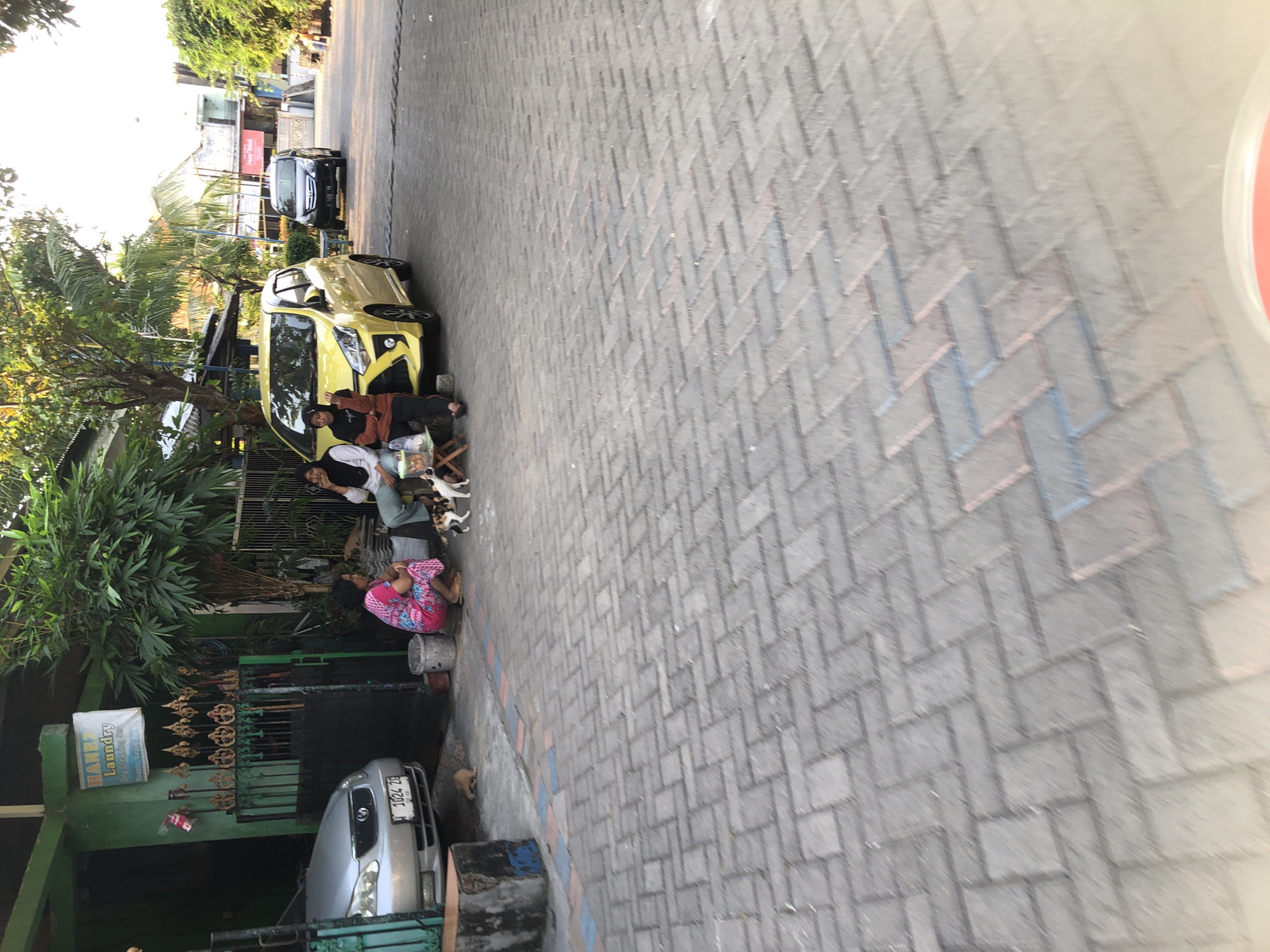 The image depicts a street scene in Kalisampurno, Indonesia, possibly in the late afternoon or early evening, as suggested by the warm, diffused light. The primary subjects are a few people gathered near a bright yellow car and a silver car parked further down the street.
In the foreground, a woman wearing a vibrant pink and blue patterned garment is seated on the ground, looking downwards. Beside her, a person in a dark top and white headscarf is seated, possibly interacting with a small dog or two, one white and one a calico. A small wooden stool is visible nearby. The yellow car appears to be parked alongside a narrow sidewalk or walkway, with a building facade and lush green foliage to its left.
Further down the street, a silver car with a license plate ending in "1024 ZE" is parked in front of a green building with a metal gate. A sign for "BANJARSARI LAUNDRY" is attached to the green building. The street itself is paved with interlocking gray bricks laid in a herringbone pattern. In the background, more buildings and trees are visible, indicating an urban or semi-urban environment. The overall impression is a quiet, everyday scene with people and vehicles on a street.