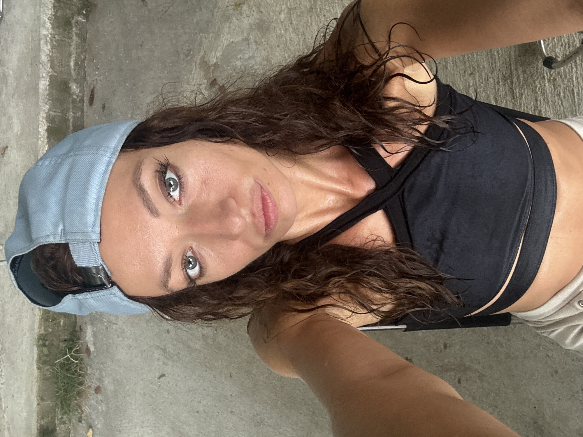 A woman is taking a selfie while standing on concrete. She is wearing a black sports bra, light-colored shorts, and a light blue baseball cap with sunglasses perched on top. Her hair is dark and appears to be wet. The background consists of a concrete surface and the bottom part of an object. There is no indication of the city or location.