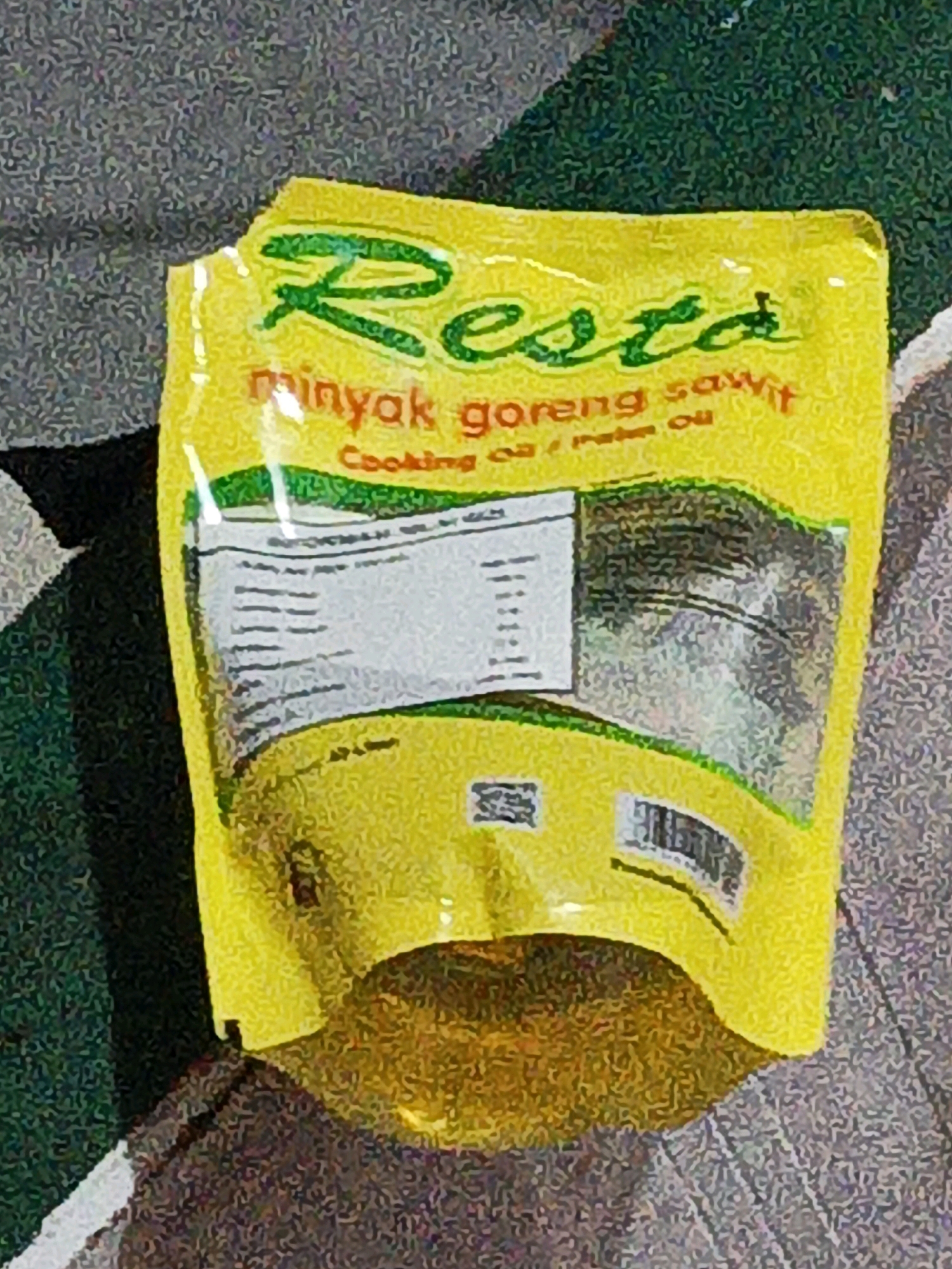 A yellow, soft plastic pouch of Resto brand cooking oil is placed on a textured, dark surface, possibly a floor or table. The pouch has green lettering that reads "Resto" at the top, followed by "minyak goreng sawit" and "Cooking oil / Palm Oil". There is a nutrition facts label on the left side of the pouch with columns of numbers and text. The pouch is partially filled with a golden-yellow liquid, presumably the cooking oil, which is visible through the translucent bottom section. The background is a blurry, speckled pattern of dark and light gray and green. The image is taken from a slightly elevated angle, looking down at the pouch. There are no people or animals visible in the image. The scene does not provide any specific clues to identify a location in West Jakarta, Indonesia, or any other city.