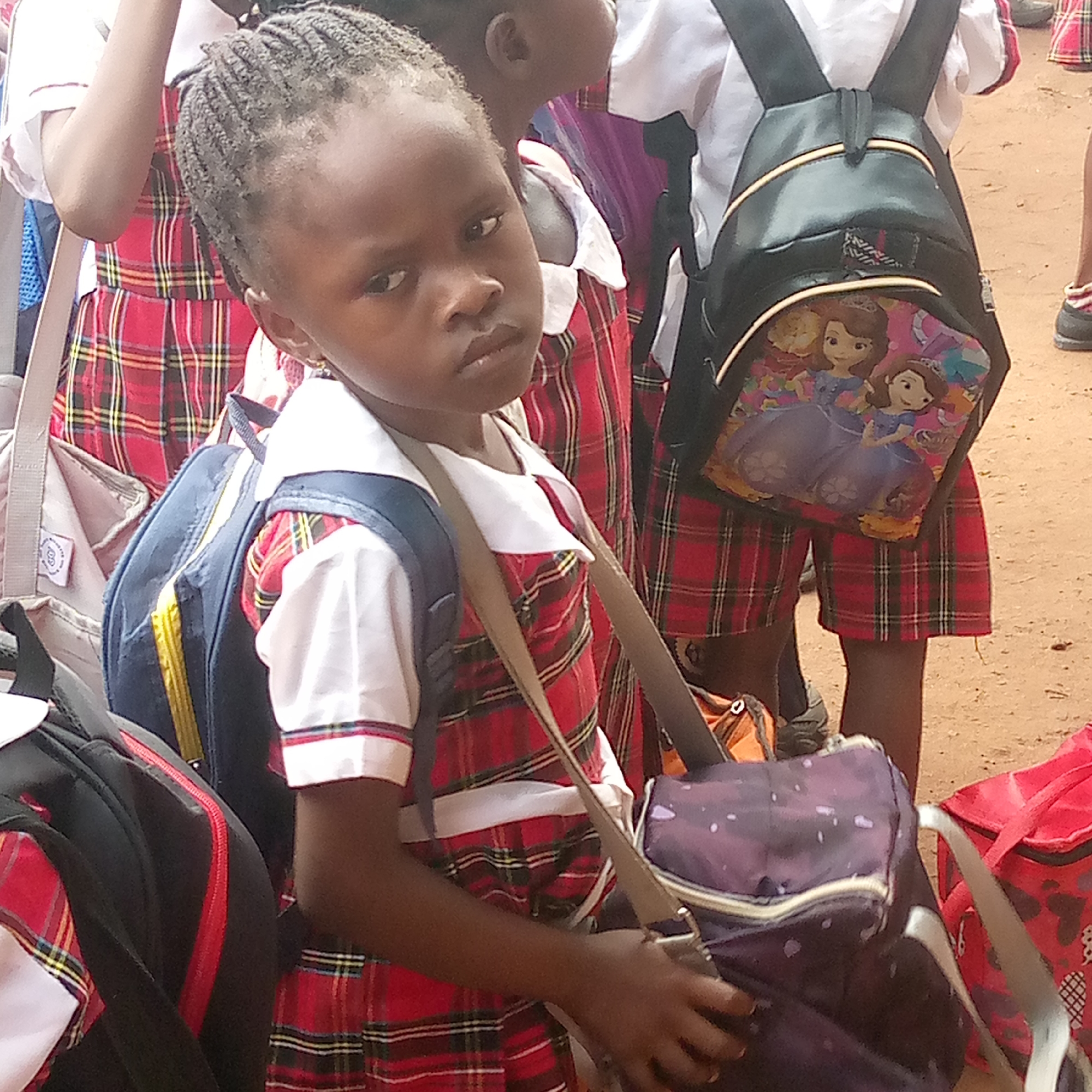 In Ukwu-Abwa, Nigeria, a group of children in red plaid school uniforms, topped with white collared shirts, are surrounded by backpacks. A young girl in the foreground has braided hair and a serious expression. She's wearing a blue and yellow backpack in addition to a purple patterned bag slung over her shoulder. Other children stand behind her, also in uniform and carrying backpacks, one of which features Disney Princesses. The environment appears to be an outdoor area with a reddish-brown ground.