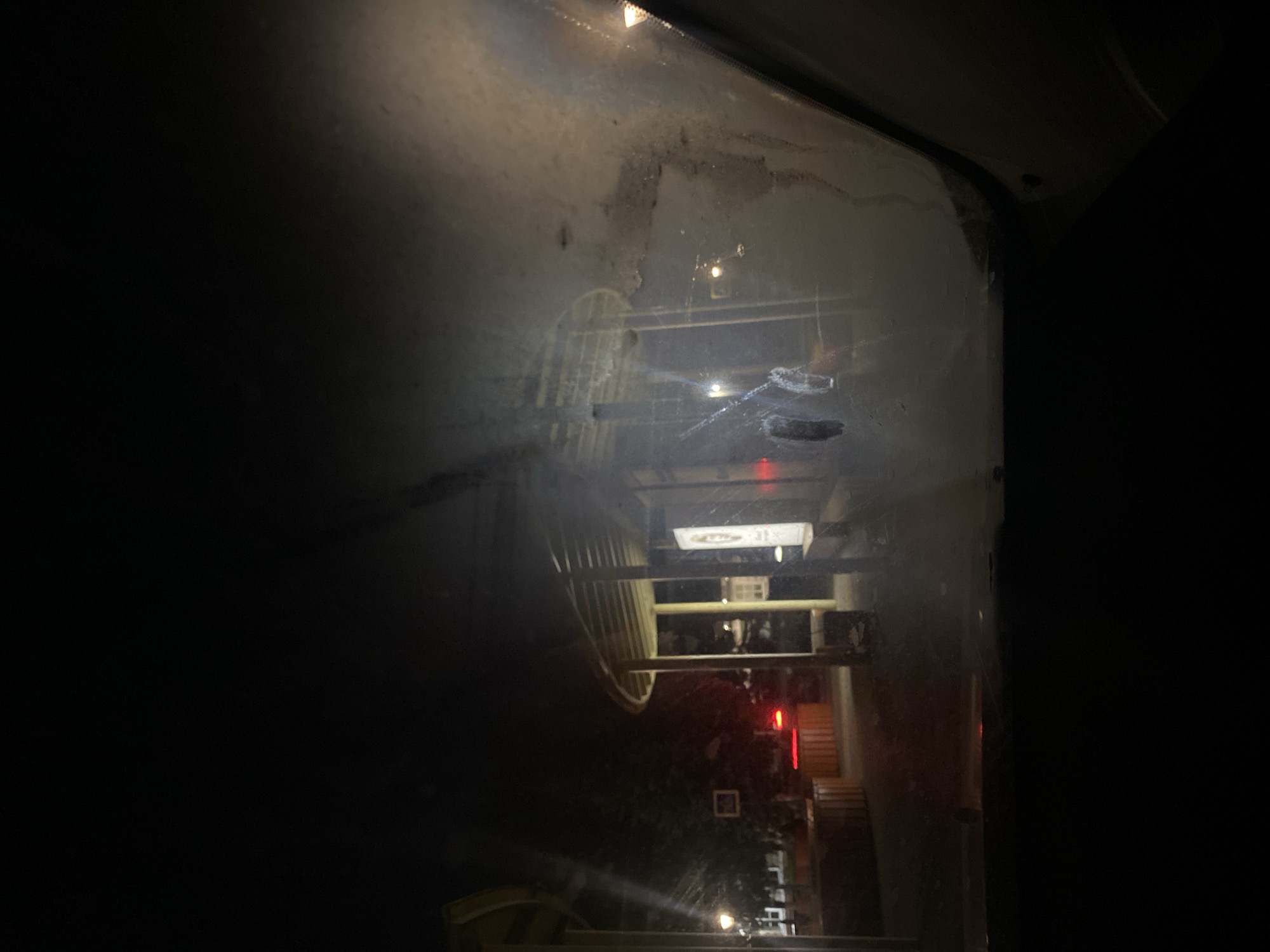 The media file presents a nighttime view looking out through a streaky, possibly dirty or fogged glass panel, which appears to be a window or a transparent barrier. The image is oriented vertically, showing reflections and smudges on the glass surface, particularly prominent in the upper-mid section, with lighter streaks and darker patches.
Through the glass, a dimly lit outdoor scene is visible. A prominent light-colored structure, possibly a railing, fence, or architectural element with numerous vertical slats or bars, is positioned towards the center-left. Beyond this structure, several artificial light sources punctuate the darkness. A distinct red, vertically oriented light source, resembling a sign or a decorative fixture, is visible in the mid-right. Above it and slightly to the left, a rectangular white light, possibly another sign, casts a soft glow. Hints of steps or tiered platforms can be discerned in the lower-mid section, leading towards the illuminated areas.
The overall setting suggests an urban or built environment, observed from a contained space, possibly a building or vehicle. There are no identifiable people or specific activities taking place. The time of day is clearly night, evidenced by the pervasive darkness and the reliance on artificial illumination. No explicit text is clearly legible on any of the visible signs or structures. The scene conveys a quiet, observational mood.