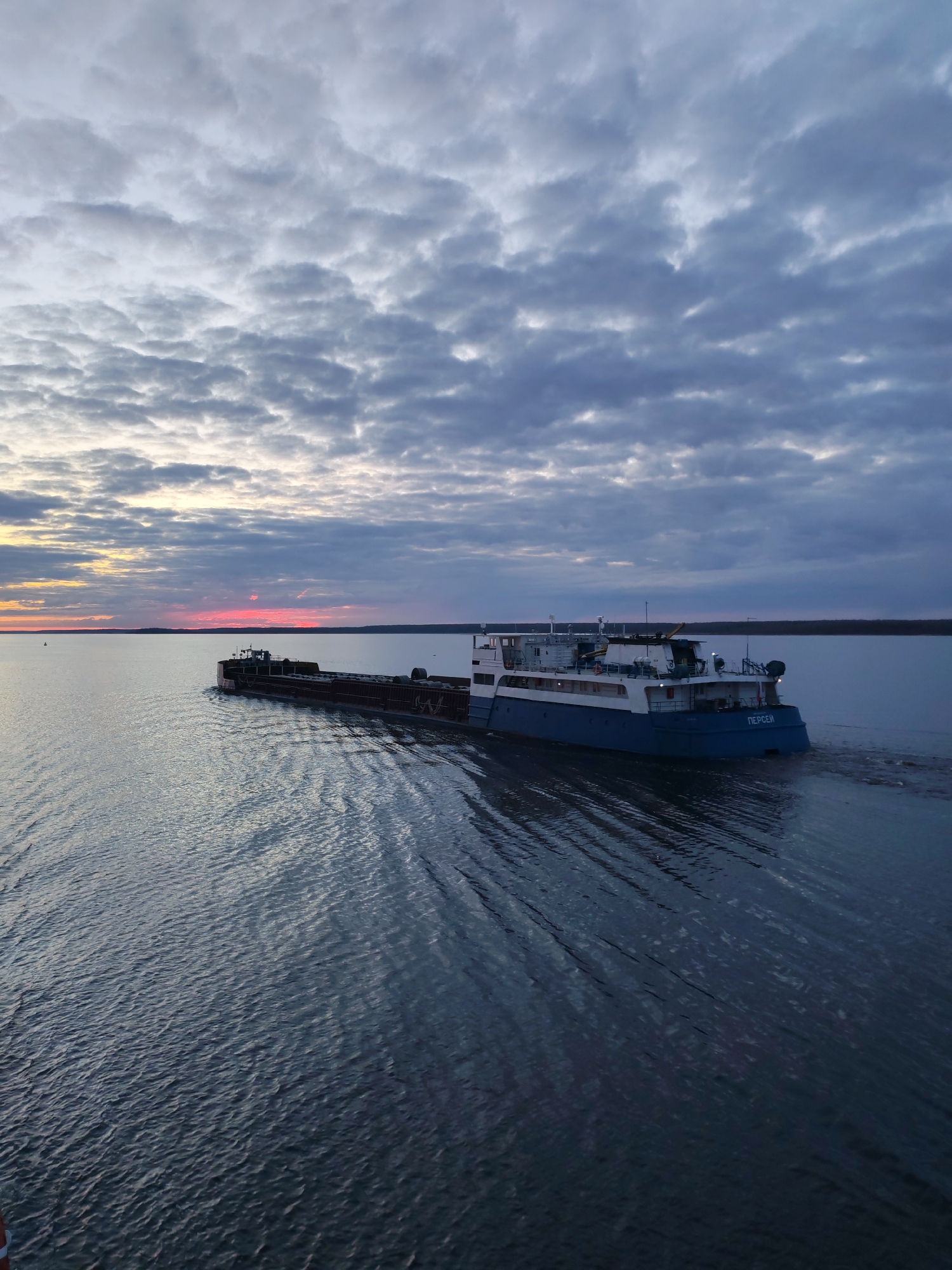 The image displays a serene twilight scene featuring a large cargo vessel navigating a wide body of water. The vessel, painted blue and white, moves from the mid-right towards the left side of the frame, leaving a distinct wake on the water's surface. On its stern, the name "ПЕРСЕЙ" (PERSEY), meaning Perseus, is clearly visible in white Cyrillic letters.
The sky is filled with numerous patchy, grey and light-blue clouds, suggesting an overcast or partly cloudy evening. On the far-left horizon, a vibrant strip of pink and orange light cuts through the clouds, indicating the setting sun. Below this colorful band, a faint, dark line of distant land or a forested shore stretches across the horizon.
The water, reflecting the muted tones of the sky, shows gentle ripples and the dynamic patterns created by the boat's movement. The overall atmosphere is tranquil, with the subtle light of dusk casting a calm yet somewhat dramatic mood. This scene captures a moment on a major waterway, likely a river or large lake, in the Kirgody region of Russia, as the vessel continues its journey during the transition from day to night.