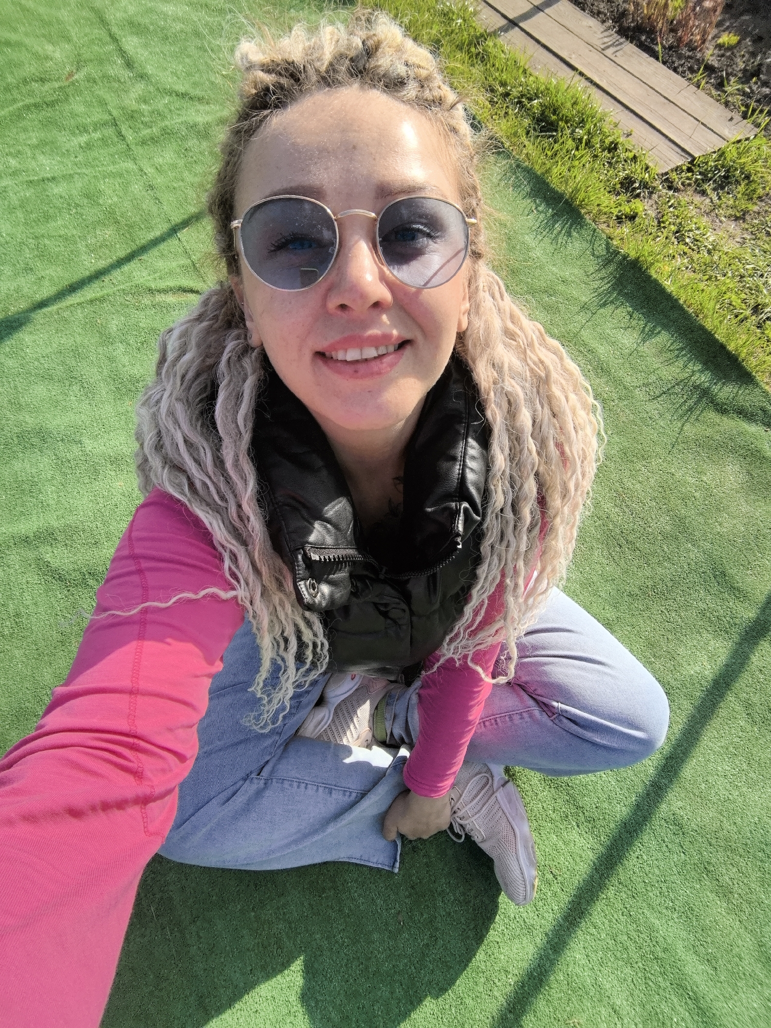 A woman with blonde dreadlocks and sunglasses is sitting cross-legged on artificial green turf, likely in a park in Noviy, Russia. She is wearing a pink long-sleeved shirt, a black vest, and light blue jeans. Her sneakers are light pink. The scene is illuminated by sunlight, casting a shadow of the woman onto the green turf. A wooden plank is visible in the background, near some grass.
