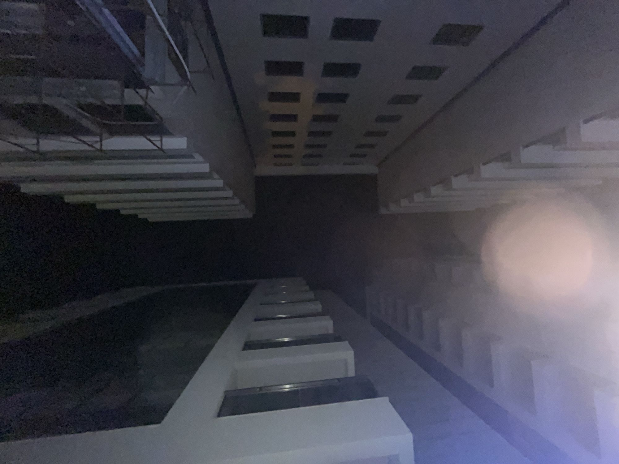 The image depicts an architectural space. At the top, there's a ceiling with square recessed lighting. Parallel walls extend downwards, with one side featuring horizontal rows of protruding structures. At the bottom, there is a wall with recessed windows. It may depict a building facade. Based on the information provided, it is in Budva, Montenegro. The overall tone is dark and obscure with lens flair obscuring the right side of the image. No people are visible.
