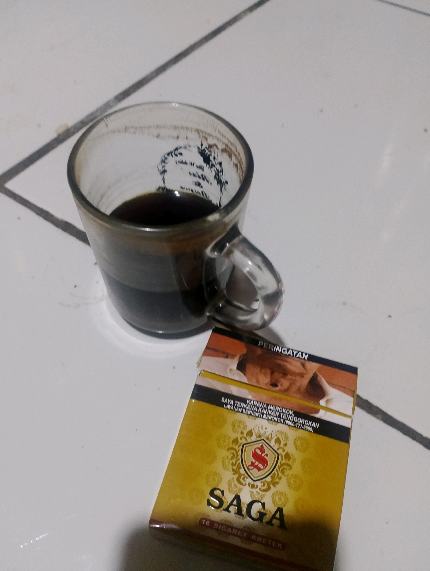 A high-angle, medium shot shows a glass of dark liquid, likely coffee, placed on a white tiled surface next to a pack of "SAGA" cigarettes. The glass has a clear handle and appears to have some residue on its interior. The cigarette pack is primarily gold and features a prominent red logo with the letter "S" inside a shield. The warning text on the pack reads "PERINGATAN KARENA MEROKOK, SAYA TERKENA KANKER TENGGOROKAN LAYANAN BERHENTI MEROKOK (0800-177-0000)". The tiled floor has visible grout lines. The lighting is dim, and there are some dust particles scattered on the surface. The image appears to be taken indoors, possibly in a residential setting in the city of Ciomas, Indonesia.