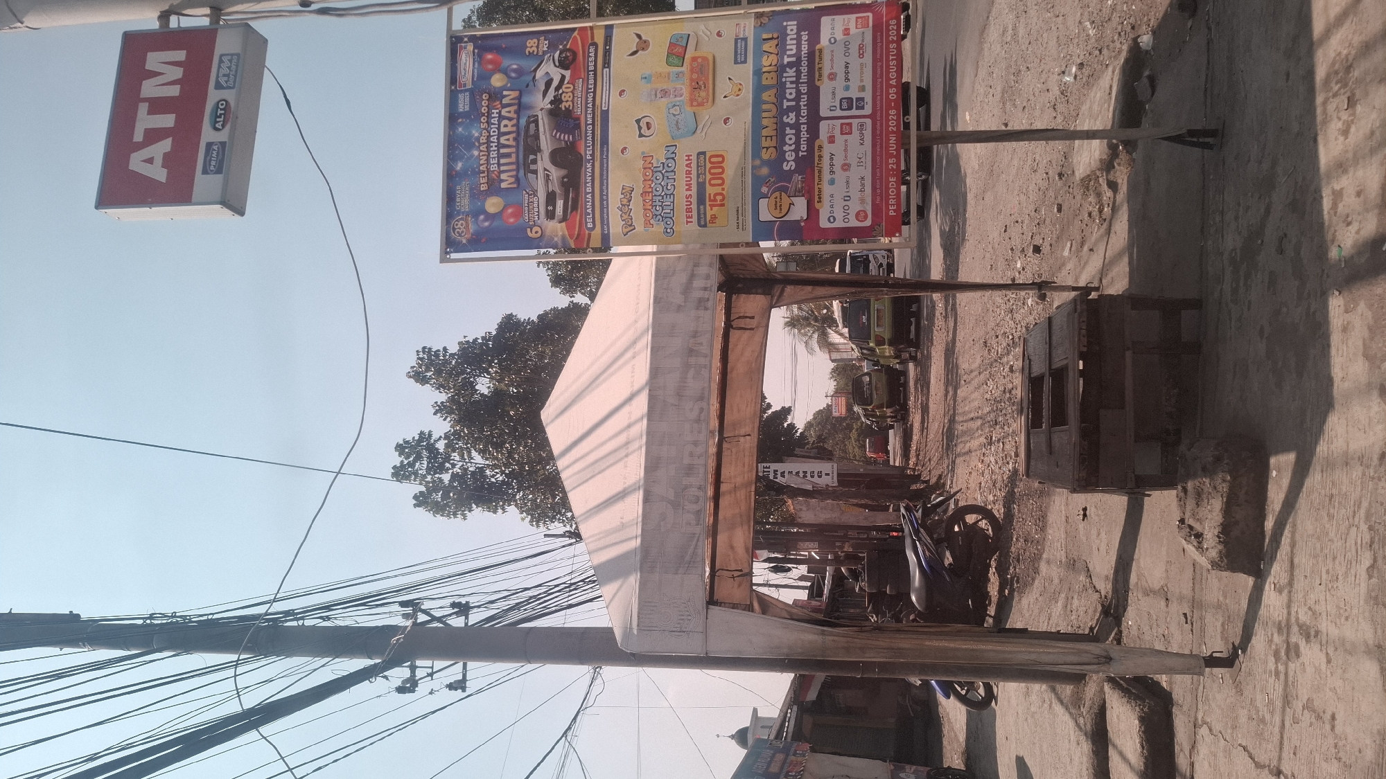 The image depicts a street scene in Rancagoong, Indonesia, under a clear sky with visible power lines overhead. In the foreground, a white tent with the words "POLRES CI..." visible on its side is set up, appearing to be a temporary stall or booth. To the left of the tent, a blue motorcycle is parked on a concrete sidewalk. In front of the tent, a wooden structure, possibly a table or a display, sits on the ground.
Further down the street, a large banner is displayed on a pole. The banner advertises promotions from Indomaret, featuring a Pokemon School Collection and a lottery with prizes in the billions. It also lists various payment methods and a period from June 25, 2026, to August 05, 2026.
On the right side of the image, a red and white ATM sign with the logos "PRIMA," "ALTO," and "ATM Bersama" is mounted on a pole. Several vehicles, including green vans and a white van, are visible on the road. Buildings with shops and a mosque with a dome can be seen in the background, suggesting a commercial and residential area. The ground is a mixture of dirt, gravel, and paved surfaces. The overall scene suggests a bustling street with commercial activity and public services.