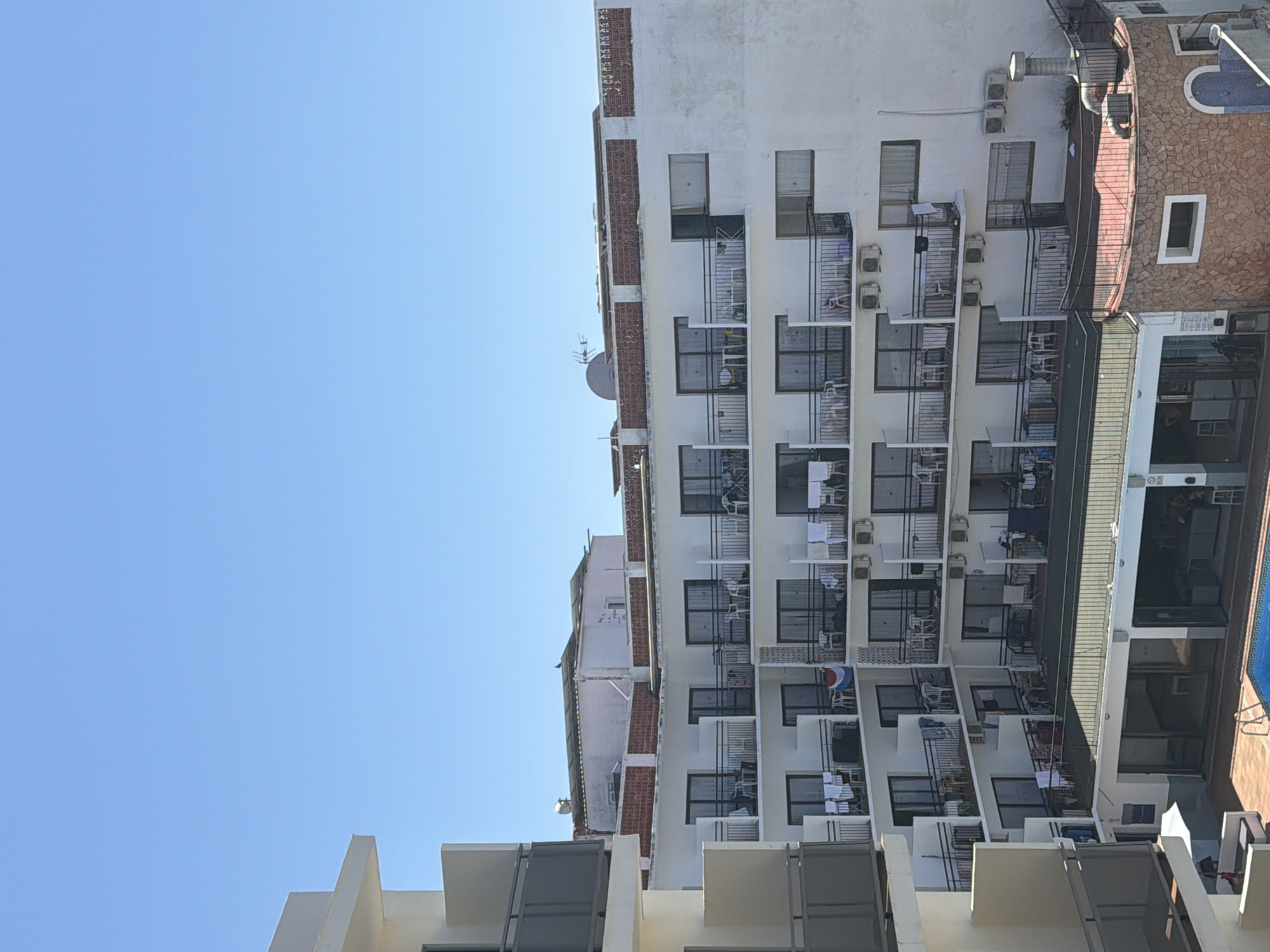 The image shows a section of a multi-story building with balconies, under a clear blue sky. The building is predominantly white with dark railings on the balconies. Various items are visible on the balconies, including chairs, tables, and laundry hanging to dry. In the foreground, there is a swimming pool and an outdoor seating area with a white umbrella. To the right, a section of the building features a rounded, stone-clad base with a blue arched doorway. The scene appears to be a hotel or apartment complex. Based on the architectural style and the presence of a pool, it is likely located in a tourist area. The city of Sant Antoni de Portmany in Spain is known for its beaches and lively atmosphere, and this type of resort architecture is common in such locations.