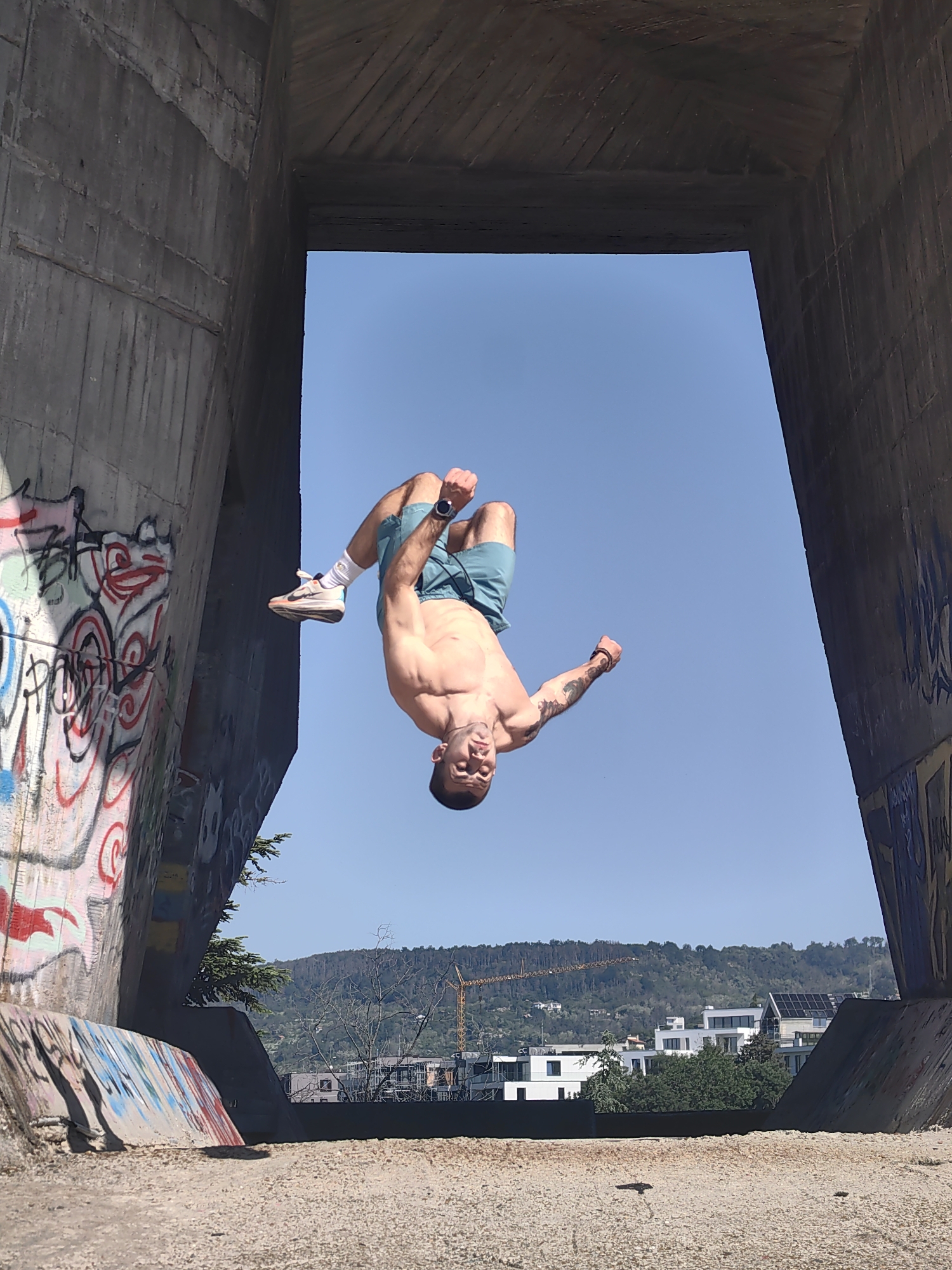 A man is doing a backflip in an urban concrete structure. The location is likely in Varna, Bulgaria, based on visual clues. The man has a muscular build, is shirtless, and is wearing blue shorts and sneakers. He is mid-air, upside down, against a clear blue sky. The concrete structure is weathered and covered in graffiti. In the background, there are buildings, trees, a hill, and a construction crane. The ground below is dirt or gravel.