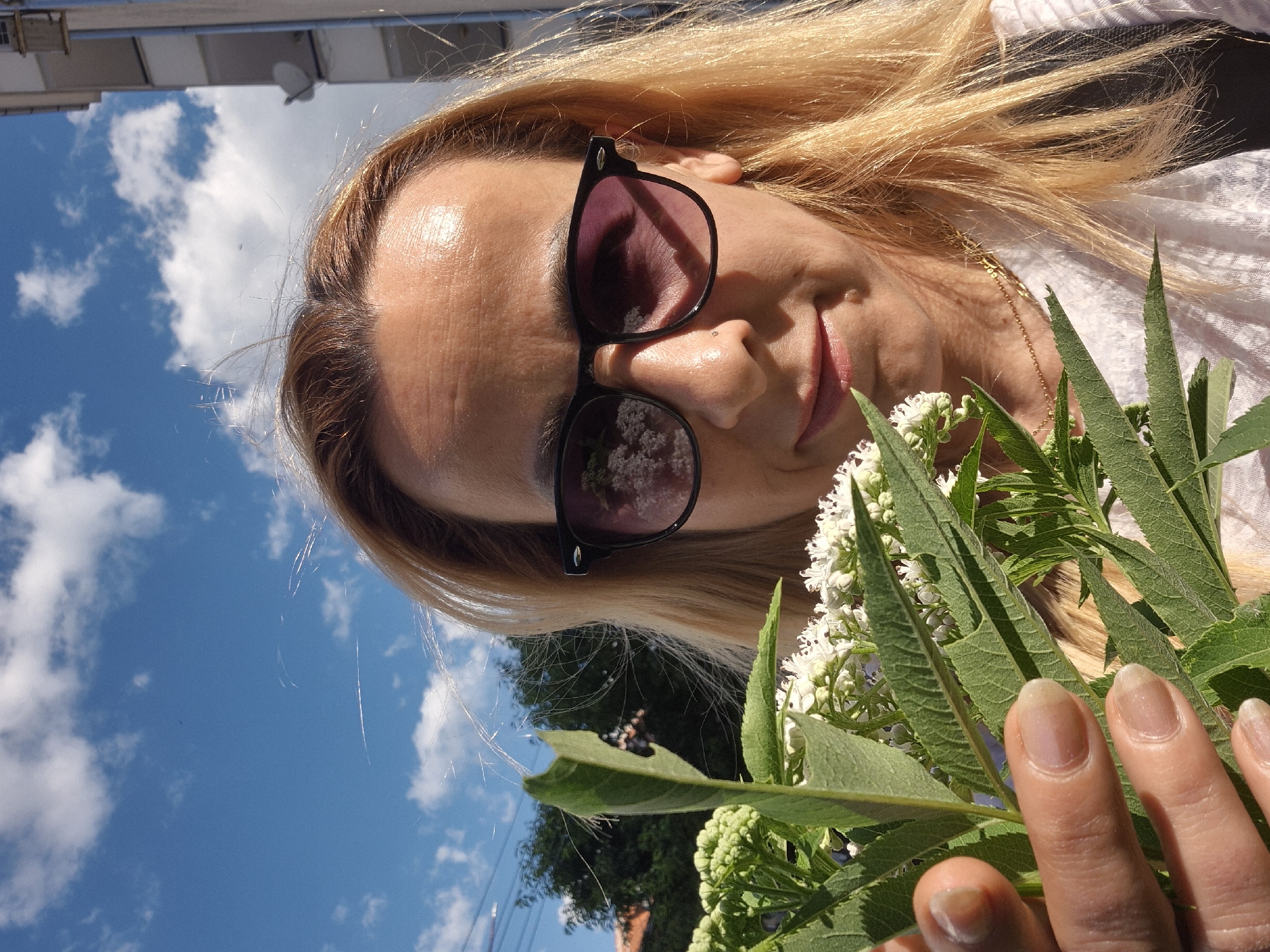 In an outdoor, urban setting, a woman with blonde hair and sunglasses holds a bouquet of white flowers and green leaves. She is positioned in the foreground, with a bright blue sky with scattered clouds behind her. A building and trees are visible in the background. The woman is wearing a white shirt and a gold necklace. Her left hand is holding the flowers, with her fingernails visible.