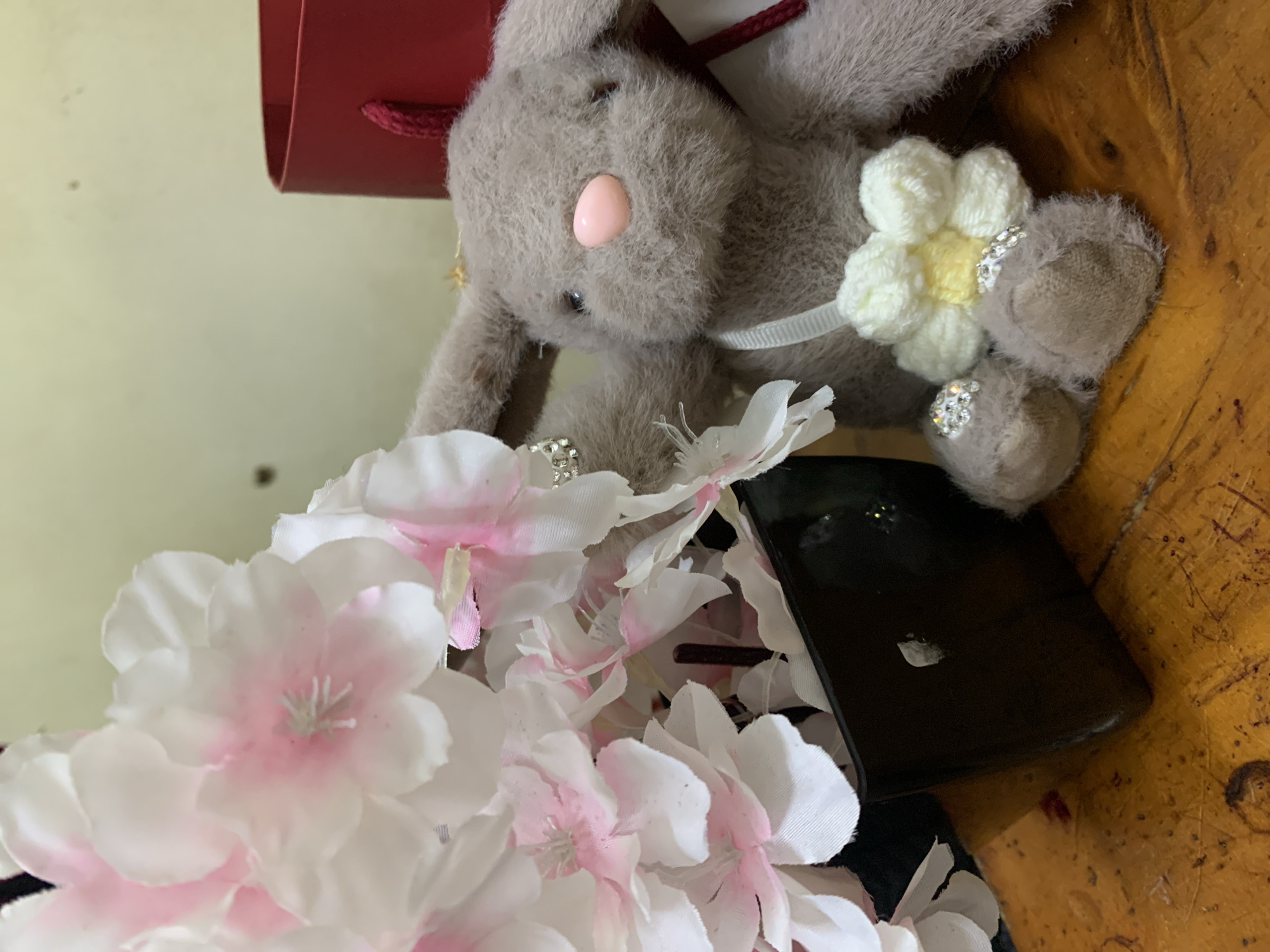A still life photograph presents a fuzzy grey bunny toy with a pink nose, adorned with a white crocheted flower and gem-like embellishments. The bunny sits atop a dark, rectangular object, possibly a phone or case, and is surrounded by artificial white and pink cherry blossoms. A red gift bag appears in the background. The setting is a textured, possibly wooden, surface. There is no indication of the location being Dar es-Salaam, Tanzania.