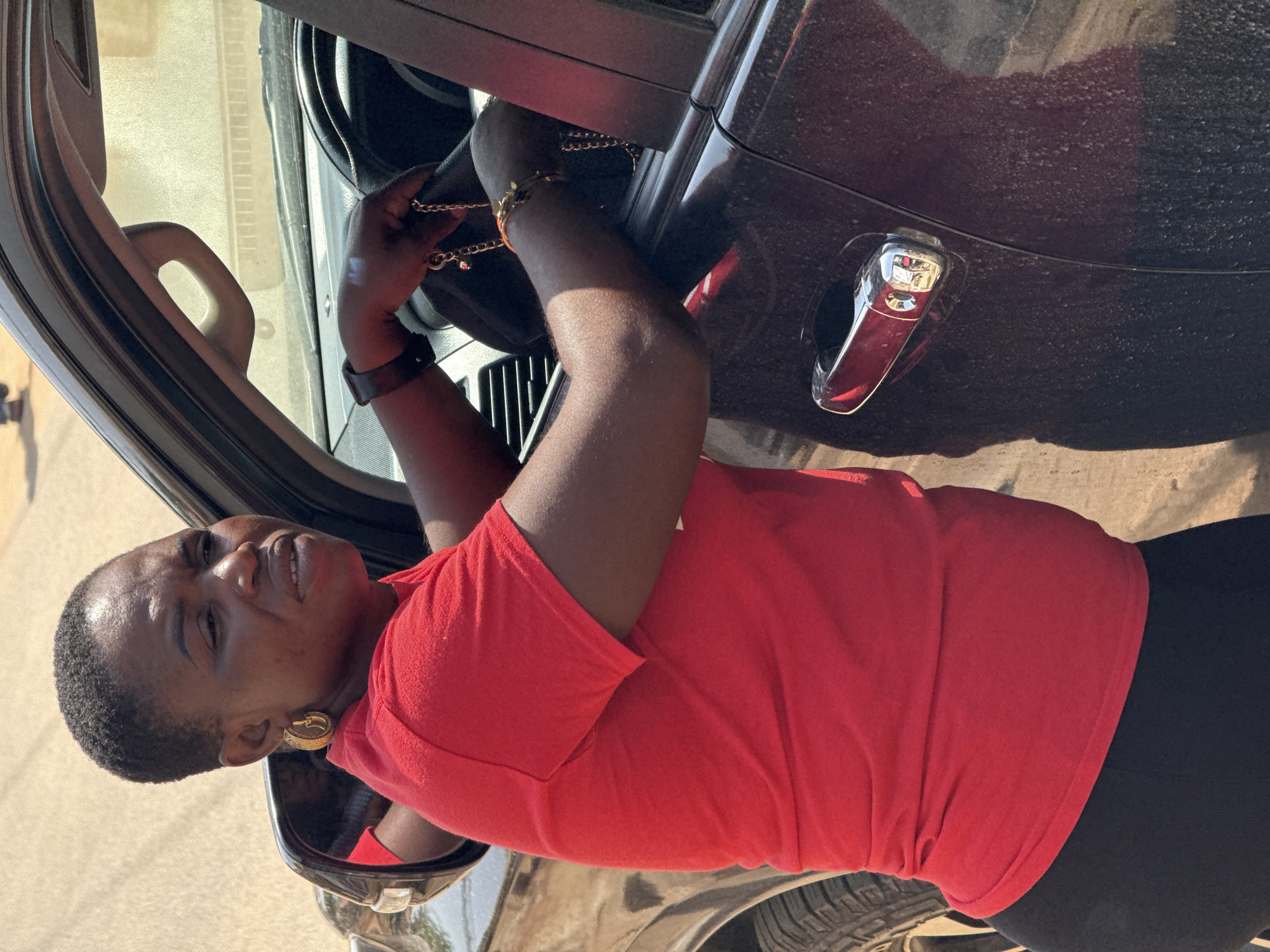 A woman with short, dark hair and wearing a bright red t-shirt and black leggings is leaning into the open driver's side door of a dark-colored SUV. She has large gold hoop earrings and is wearing a watch on her left wrist. Her hands are inside the car, and she appears to be holding a chain or necklace. The car's interior is visible, including part of the steering wheel and dashboard. The scene appears to be outdoors, with a dusty, light-colored ground and a hint of buildings or a wall in the background. The lighting suggests it is daytime, possibly a sunny day. The image was taken in Abuja, Nigeria.
