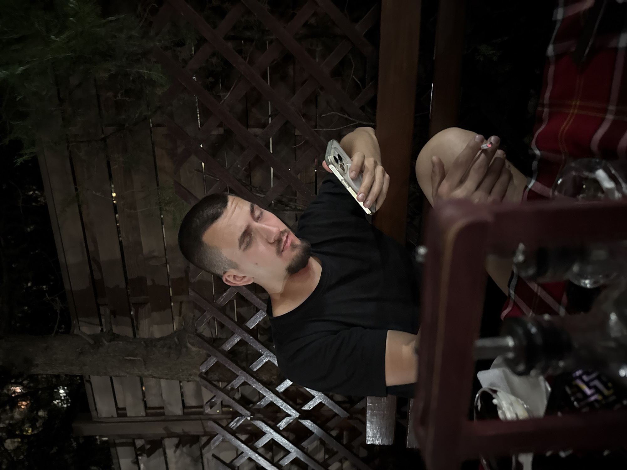 A man with a short haircut and a goatee is seated outdoors at night, possibly in Sofia, Bulgaria. He holds a smartphone in his left hand and a lit cigarette in his right hand. He is wearing a black t-shirt. A red and white plaid pattern is visible in the upper right corner, possibly a blanket or a garment worn by another person. The setting appears to be a patio or deck, with a dark wooden trellis and vegetation visible in the background. There are glasses and other objects on a table in the foreground. The image is rotated.