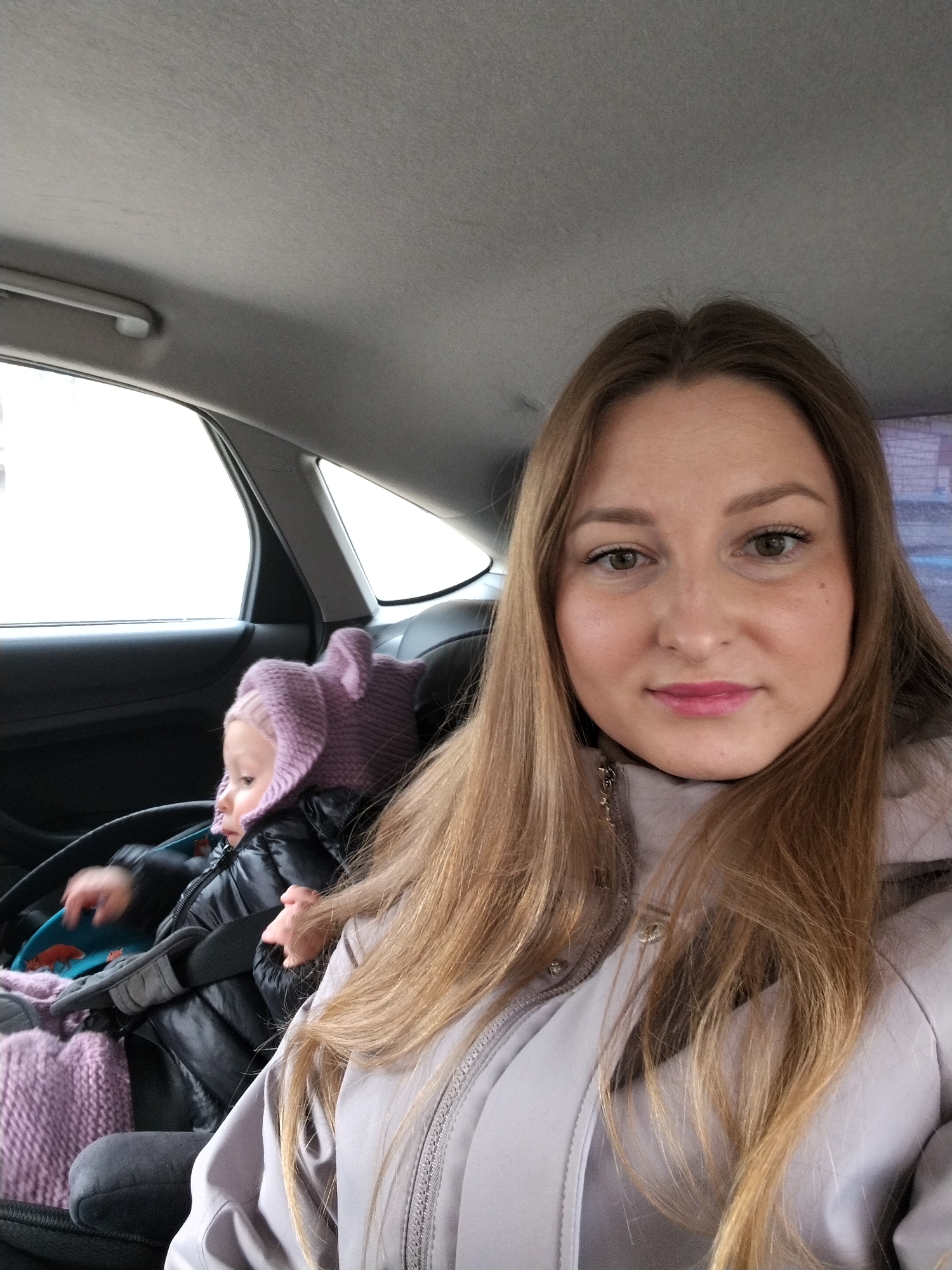 A woman and a child are in the back seat of a car. The woman has long brown hair and is wearing a light gray jacket. The child is wearing a pink hat and black puffer jacket and is secured in a car seat. The car is moving, and the scene suggests the woman and child are in Chaikovsky, Russia.