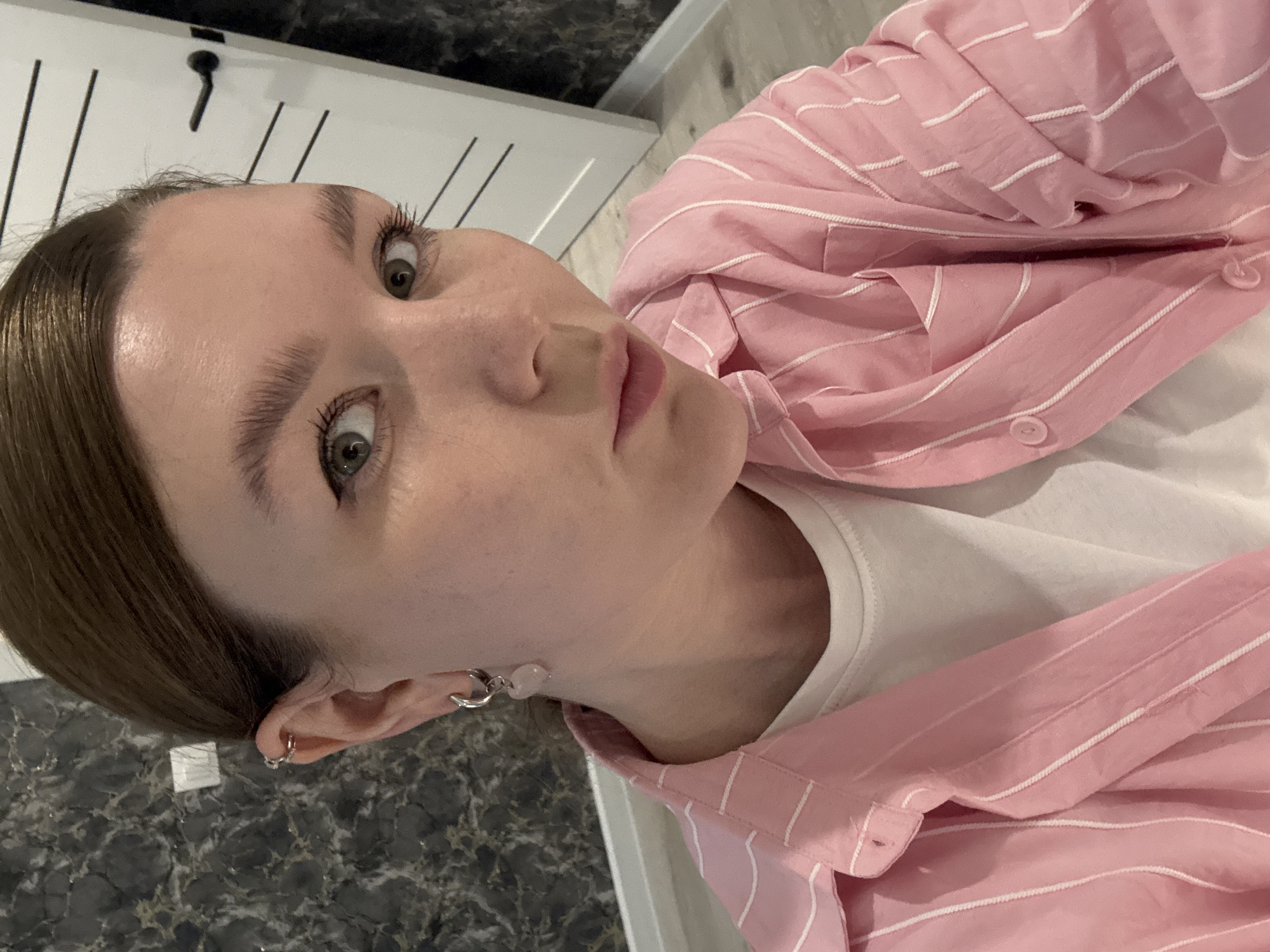 A young woman is captured in a close-up, tilted portrait, likely a selfie. She has light skin, brown hair pulled back from her face, and striking light-colored eyes with long, dark eyelashes. Her gaze is direct and neutral as she looks towards the viewer. She is wearing a light pink, collared button-up shirt with fine white horizontal stripes, open over a simple white t-shirt.
Notable details include multiple silver ear piercings: a small hoop in her helix and a larger, decorative dangle earring with a light pink or white stone on her earlobe, as well as a silver hoop visible on her other ear. The background indicates an indoor setting. To her left and below, a dark, glossy surface with a marble or granite-like pattern is visible. Behind her head and shoulder, a white wall features thin black vertical lines and a single black hook or knob. The lighting is ambient indoor light, and no specific time of day or weather can be discerned. There is no visible text in the image. The location context is Izhevsk, Russia.