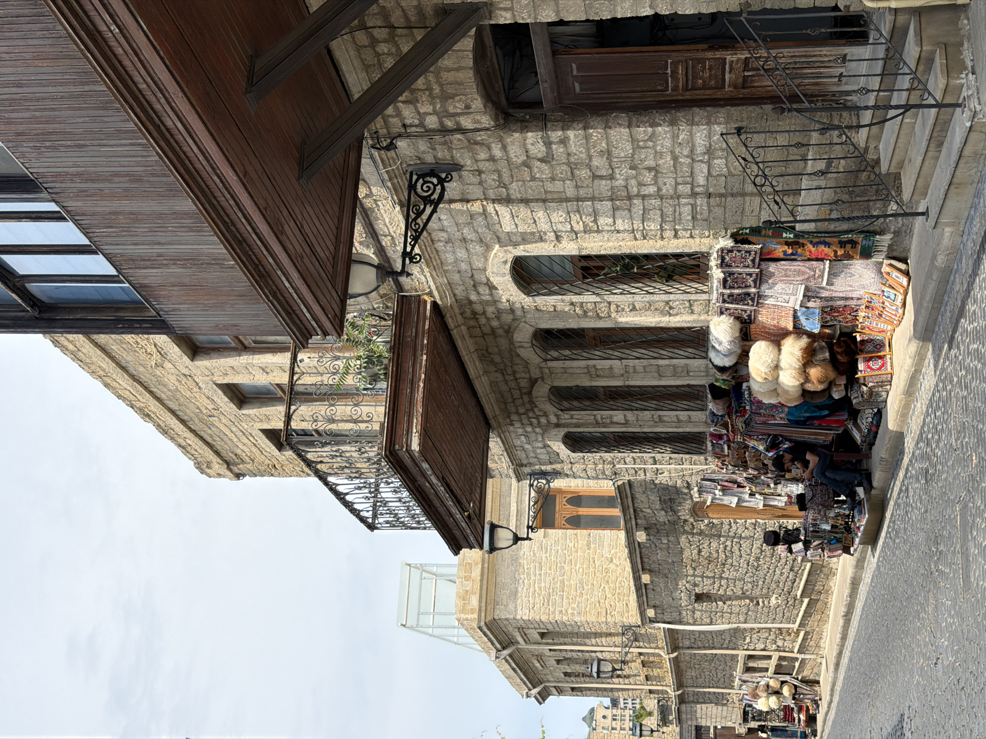 The image depicts a cobblestone street in the Old City of Baku, Azerbaijan. On the right side of the street, a two-story building constructed of light-colored stone features a wooden balcony and a wooden door. To the left of the door, a display of carpets and faux fur items is set up on the sidewalk. A person is seated on a small stool, looking at a mobile phone, amongst the displayed goods. Further down the street, more vendors are visible, with their merchandise displayed along the stone wall of another building. The street is narrow and appears to be in a historic district, with the architecture indicative of traditional Azerbaijani style. The sky is overcast, and the lighting suggests it is daytime.