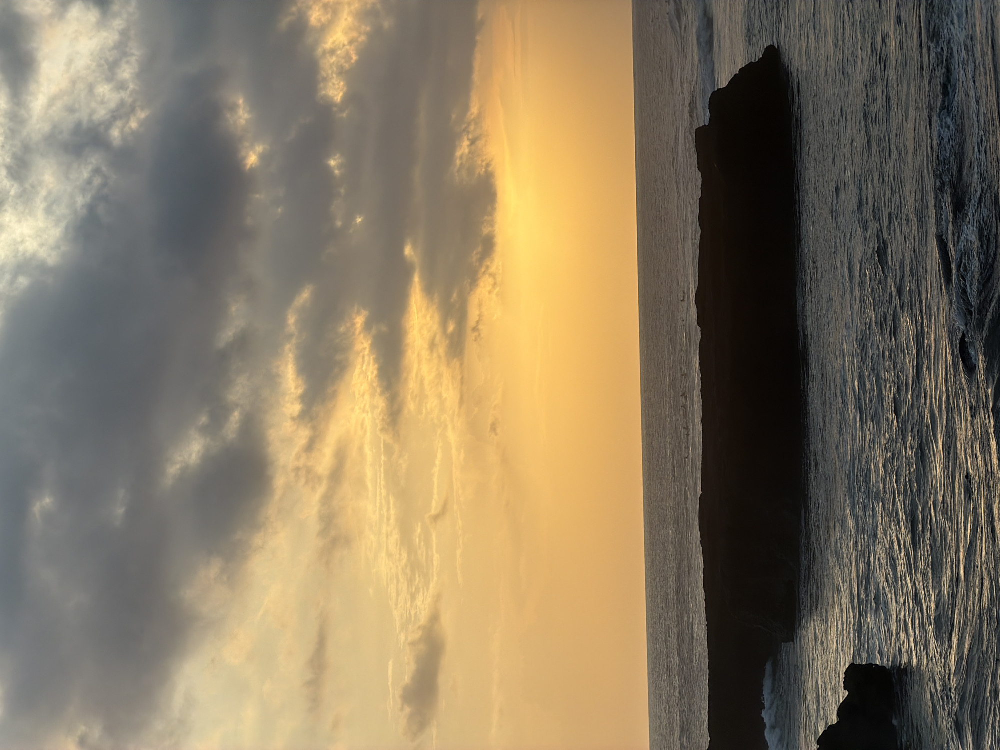 The image depicts a sunset scene in North Kuta, Indonesia, showcasing a sky filled with clouds tinged with orange and gold hues from the setting sun. The left side of the image captures the dynamic cloud formations, ranging from darker, brooding grey clouds to lighter, golden-edged ones reflecting the sunlight. The sun itself, though not directly visible, casts a warm, radiant glow across the sky, suggesting it is setting just below the horizon.
On the right side, the ocean is visible, characterized by a rocky outcrop. The ocean's surface is textured with waves, their patterns highlighted by the light, creating a stark contrast between the dark rock and the shimmering water. The overall composition of the image evokes a sense of tranquility and natural beauty, typical of coastal sunsets.