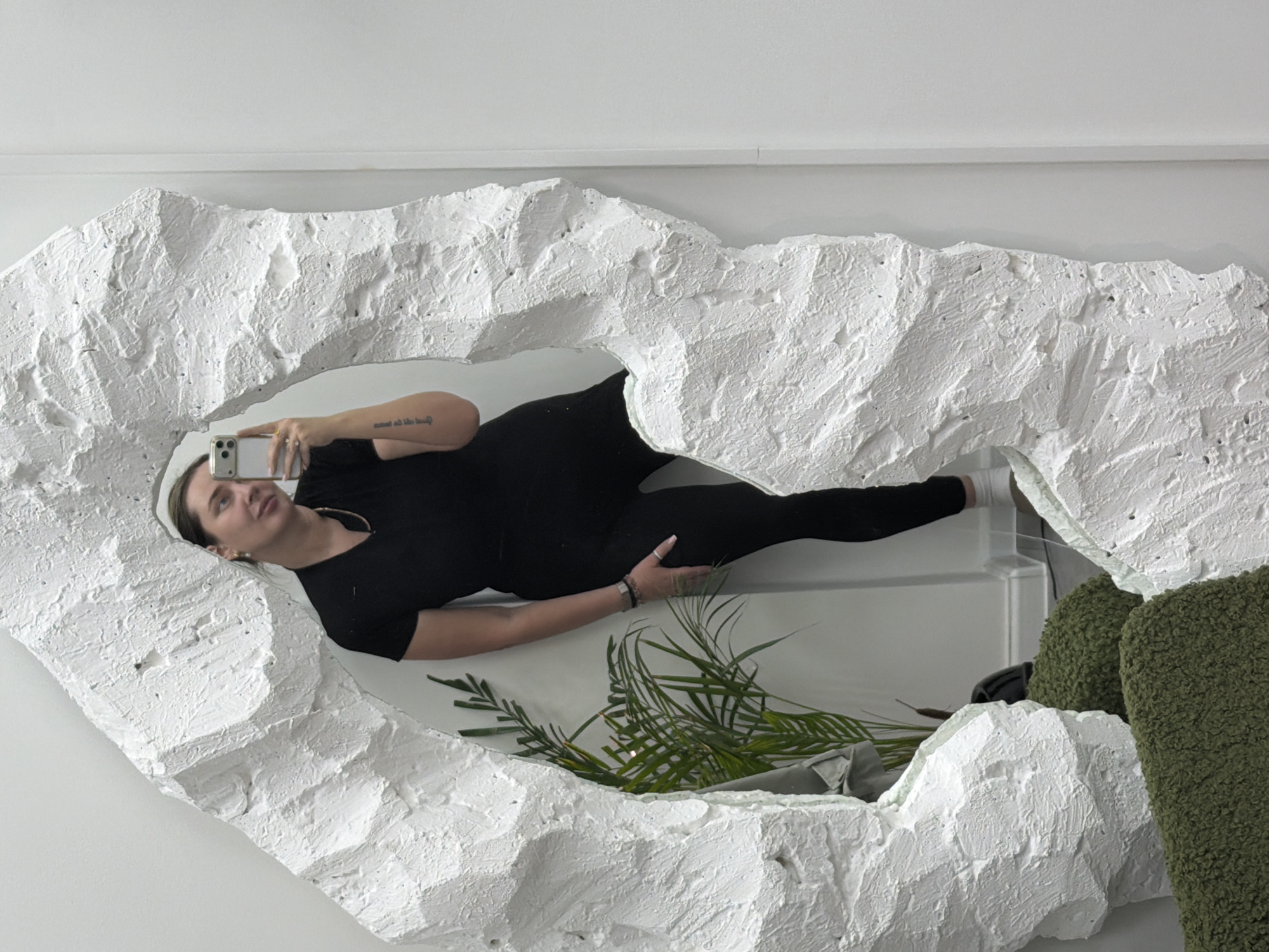 A woman in black leggings and a black t-shirt lies on her side in front of a mirror. She holds a white smartphone in her hand, ostensibly taking a picture. The mirror has an irregular, organic shape, with a rough white textured frame. A plant and a textured green object are also visible in the mirror's reflection. The location and city are not discernible from the image.