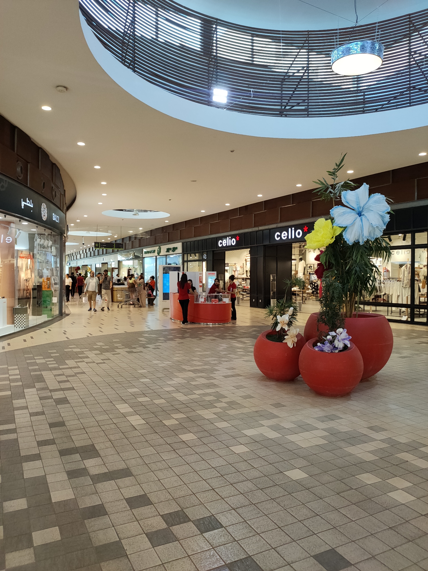 This media file shows the interior of a shopping mall in Cebalat, Tunisia. The scene is well-lit, suggesting it is daytime, and the atmosphere appears calm and typical of a shopping center.
The subjects include several people walking through the mall. Some are browsing, while others appear to be heading somewhere specific. The focus of the foreground is a decorative arrangement of three large, red, spherical planters containing artificial plants and large, colorful fabric flowers. Two of these planters hold bamboo-like foliage with large blue and yellow flowers, and the smaller one has delicate white and purple flowers.
In the background, various shops are visible along the mall corridor. Brands like "celio*" are prominently displayed on the storefronts, indicating clothing retailers. To the left, a shop with a black facade and a sign with Arabic script and "BALTI" is also present. Several glass-fronted displays showcase merchandise. People are seen entering and exiting stores, and a few individuals are gathered near a red counter, possibly at a service desk or information point. One person in a red shirt is interacting with a customer at this counter.
The architecture of the mall includes a distinctive curved ceiling with a modern, slatted design, and a large, circular light fixture hangs from it. The floor is tiled with a patterned arrangement of grey and beige squares, creating a mosaic effect. The overall impression is of a clean, modern commercial space designed for shopping and leisure. No specific events or emotional expressions are particularly highlighted, suggesting a routine day at the mall.