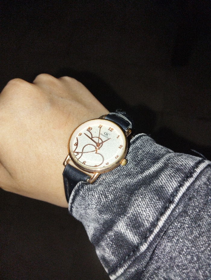 A person's left arm, adorned with a round wristwatch featuring a white face, black band, and rose gold casing. A heart design is visible on the watch face, accompanied by numerals marking the hours. The arm is covered by a grey denim or similar textured fabric jacket or shirt sleeve. The background is dark and indistinct.