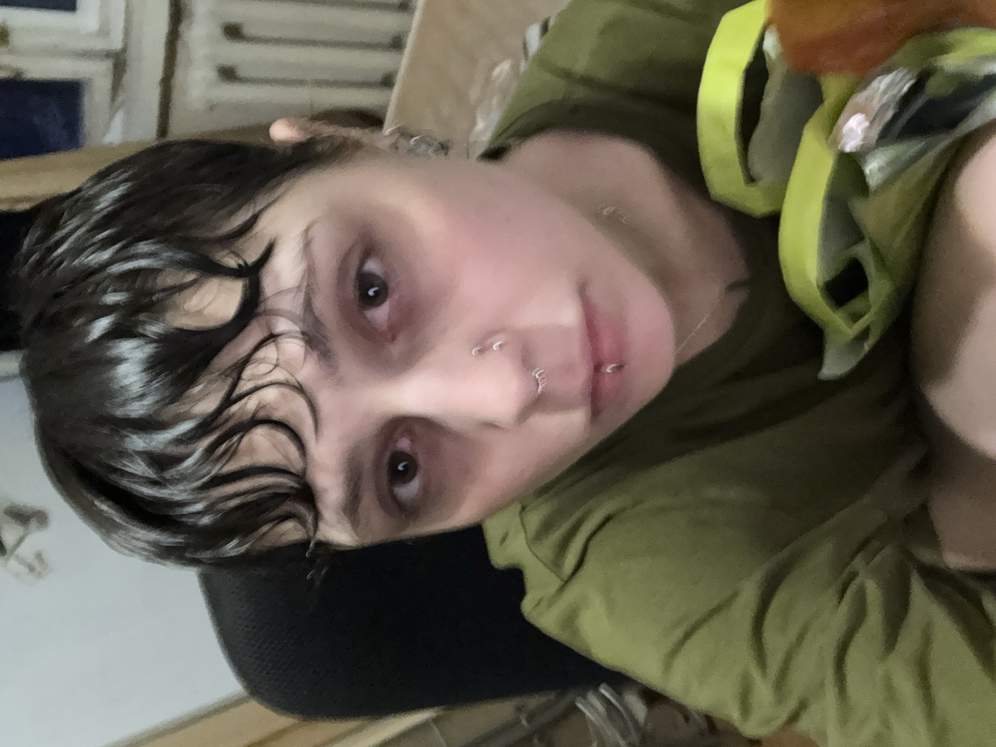 A selfie perspective features a person with dark, wet hair styled in waves across their forehead. They have brown eyes, fair skin, and multiple piercings on their nose and lip. The person is wearing an olive green shirt. The background is indistinct but suggests an indoor setting with furniture and some light coming from the left side. There is no evidence to suggest this is in Moscow, Russia.
