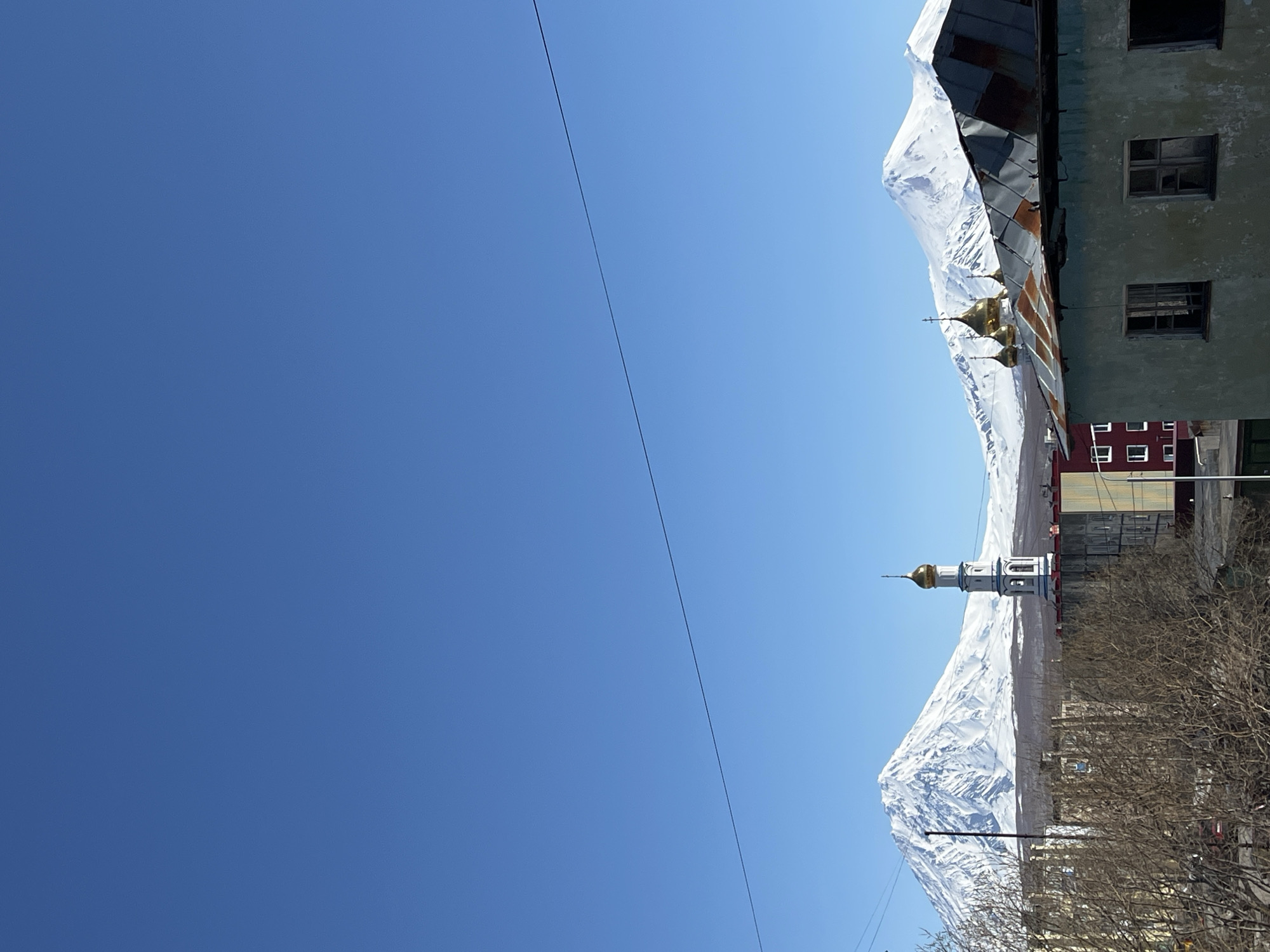 The image is oriented vertically, showing a view of Petropavlovsk-Kamchatsky, Russia. The sky is clear and blue, taking up the majority of the left side of the frame. A thin black power line runs diagonally across the sky. In the background, partially obscured by bare trees, is a white church with a golden dome, nestled in front of a snow-covered mountain. Adjacent to the mountain, a weathered building with visible windows is present. The overall scene depicts an urban environment with natural elements.